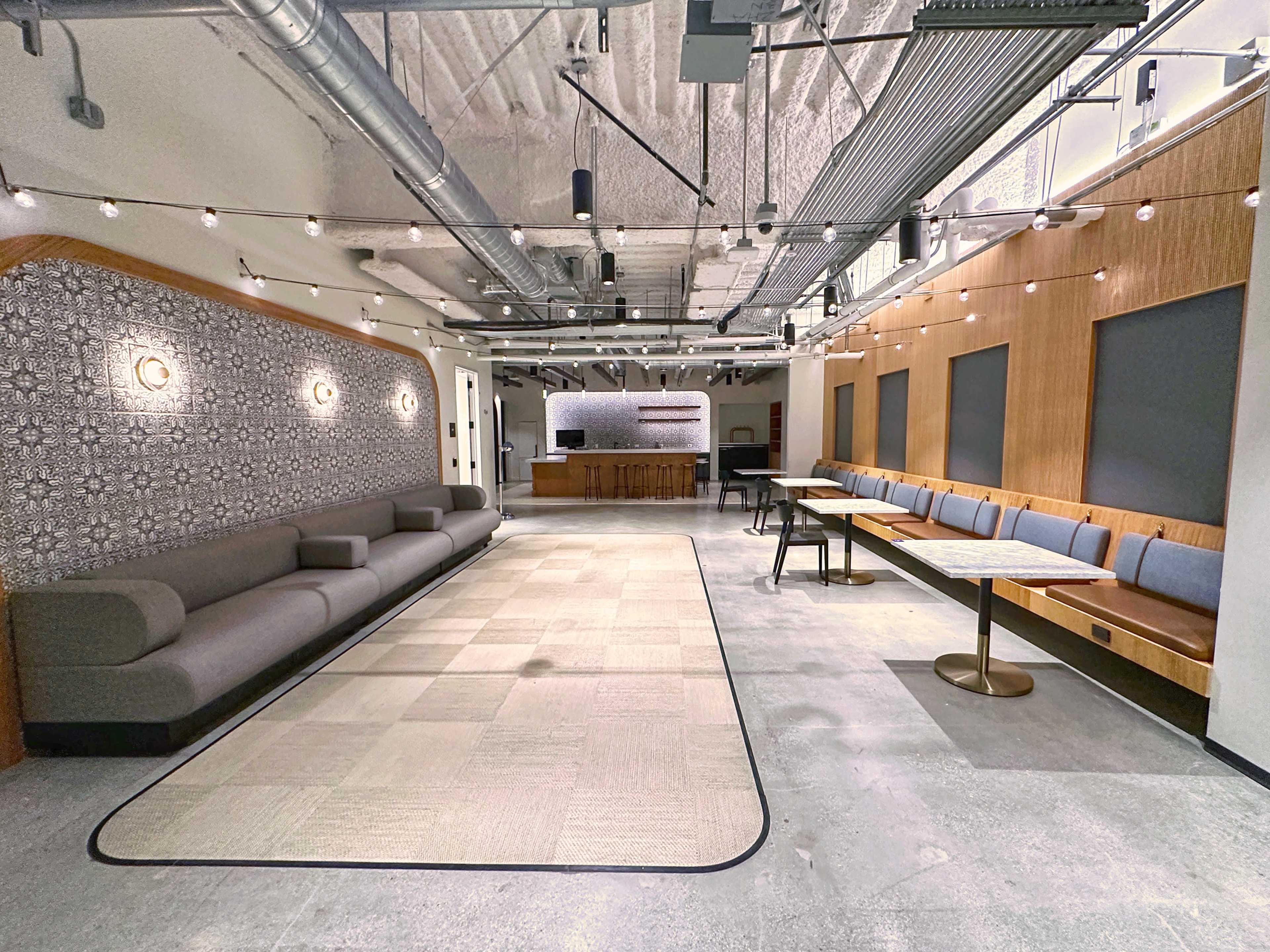A spacious interior featuring a long gray sofa, a series of tables with wooden benches, and decorative lighting overhead.