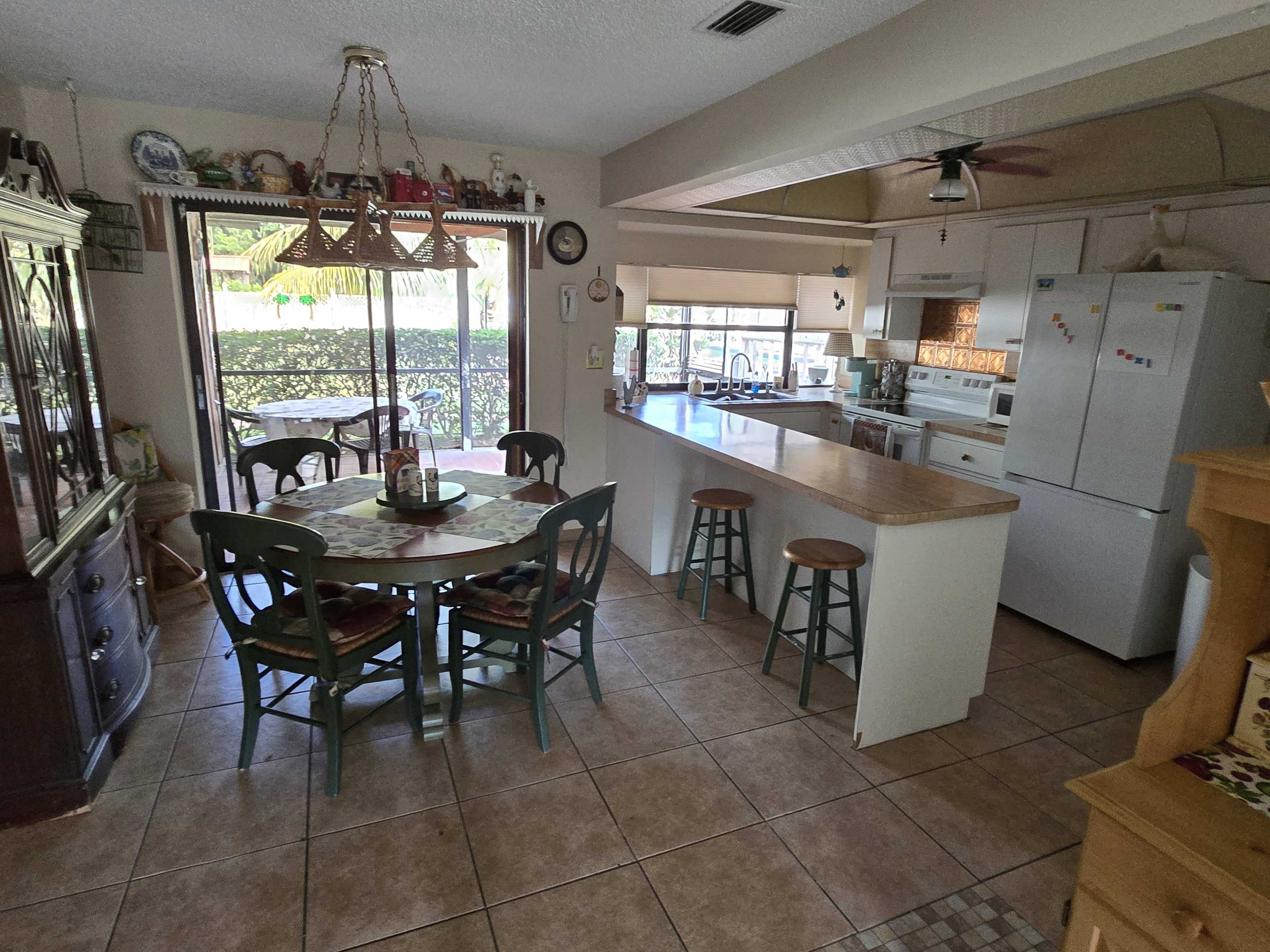 The image shows a kitchen and dining area with wooden furniture, a large table, and an open layout that connects to an outdoor space.