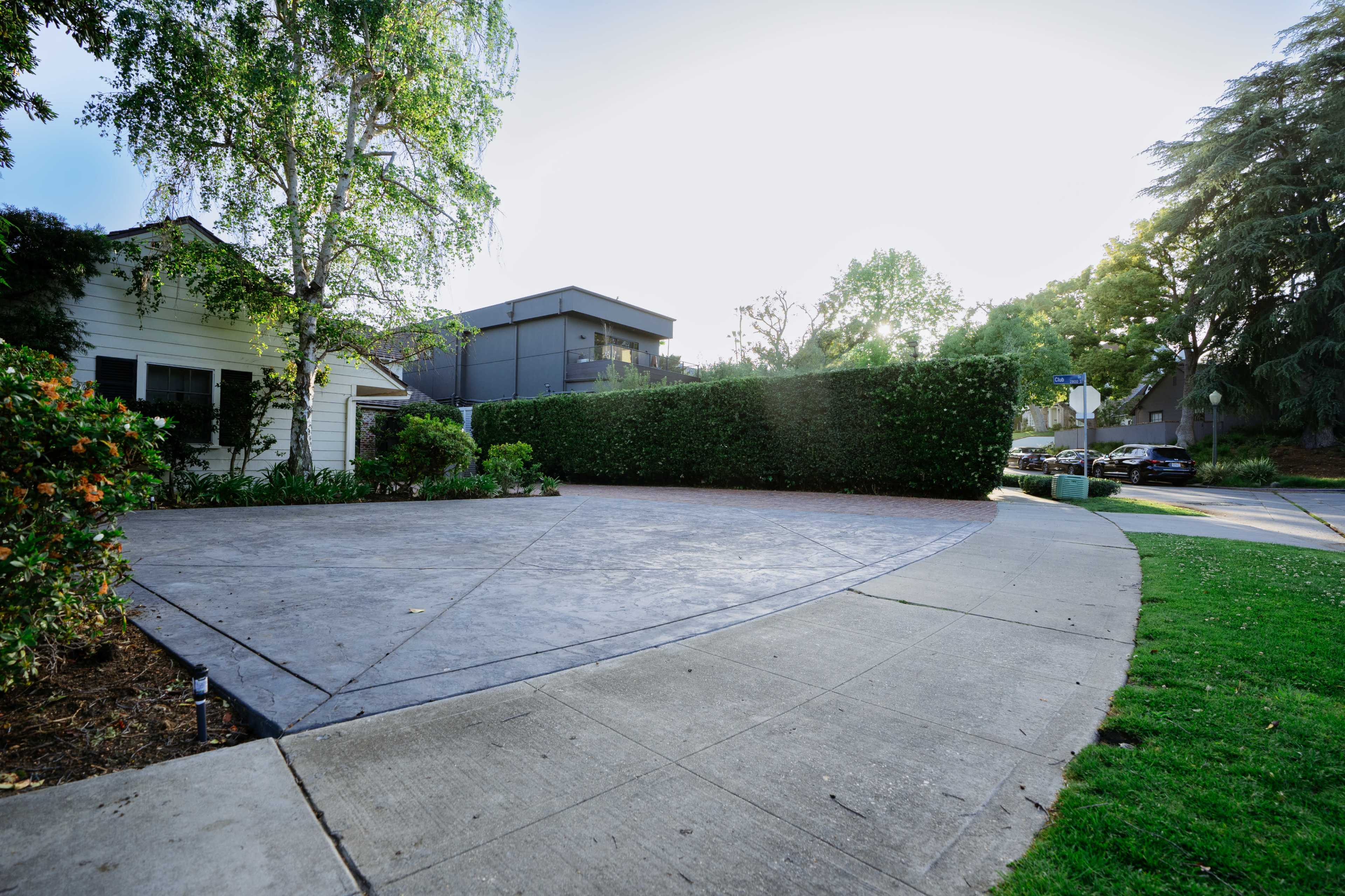 The image shows a curved driveway leading up to a house, bordered by a hedge and a sidewalk.