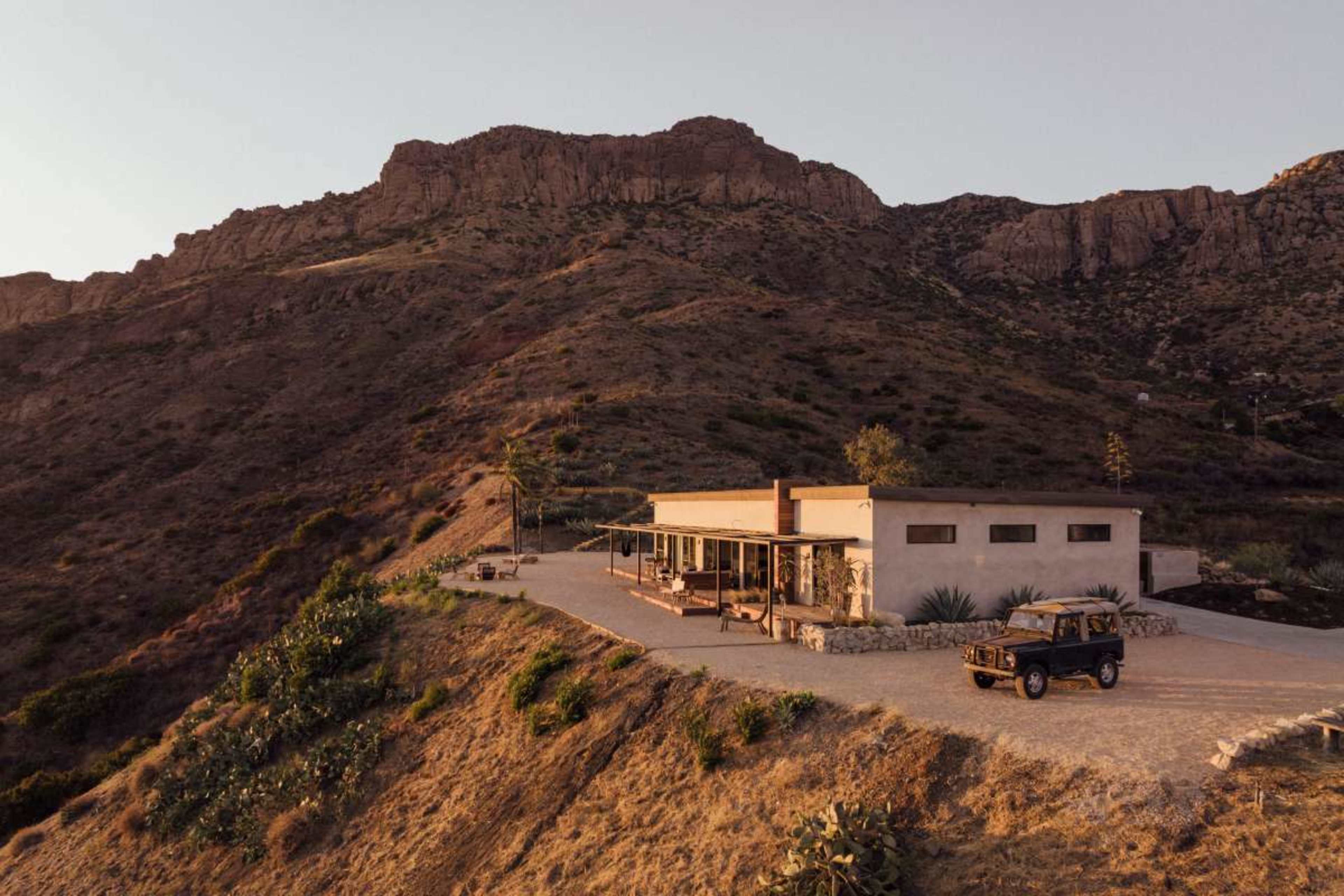 A modern house sits on a hillside surrounded by rocky terrain and sparse vegetation, with a vintage jeep parked nearby.