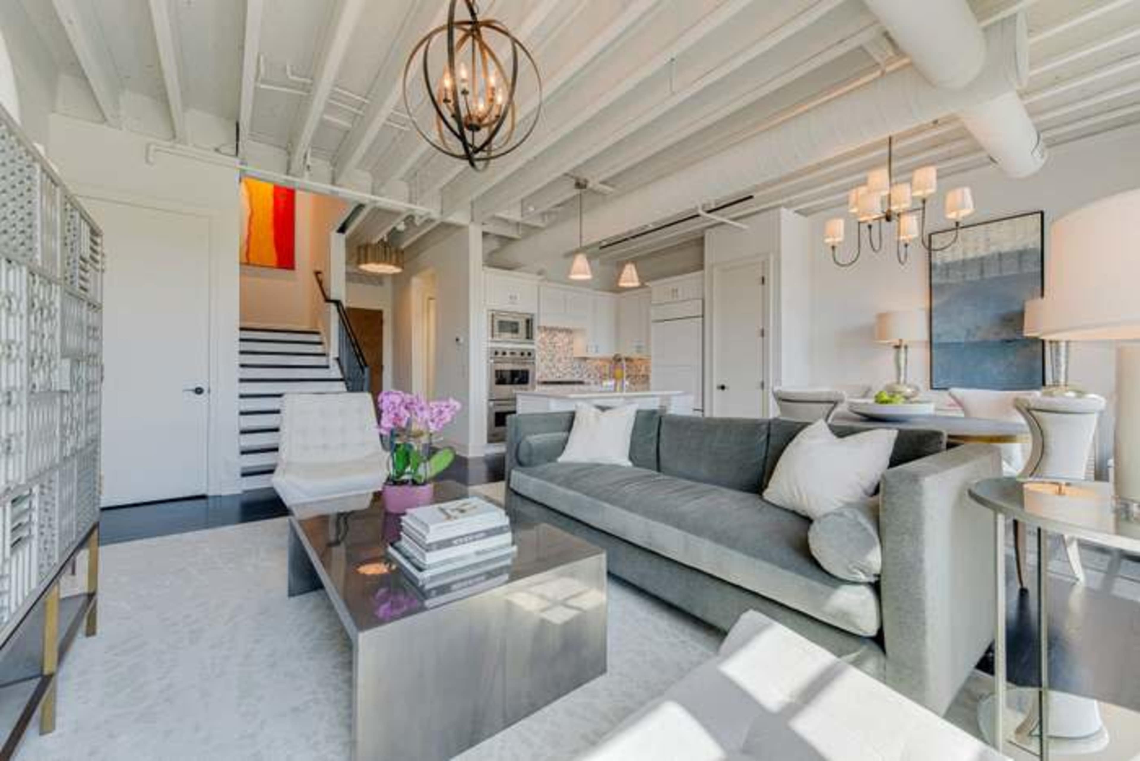 The image shows a modern living room featuring a gray sofa, a glass coffee table, and a staircase leading to another level.