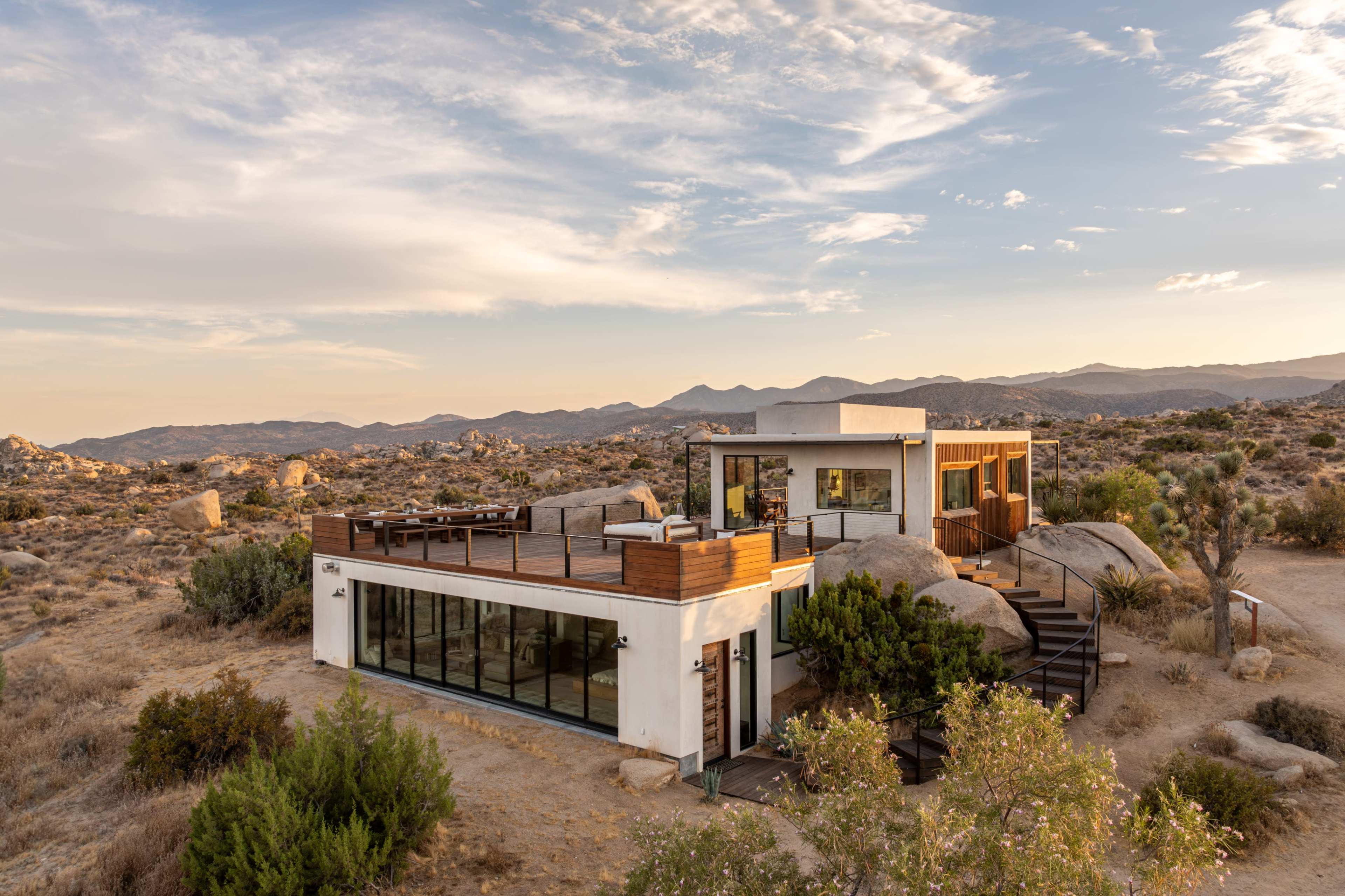 A modern house with large windows is situated among boulders in a desert landscape, with mountains in the background and a wooden deck in front.