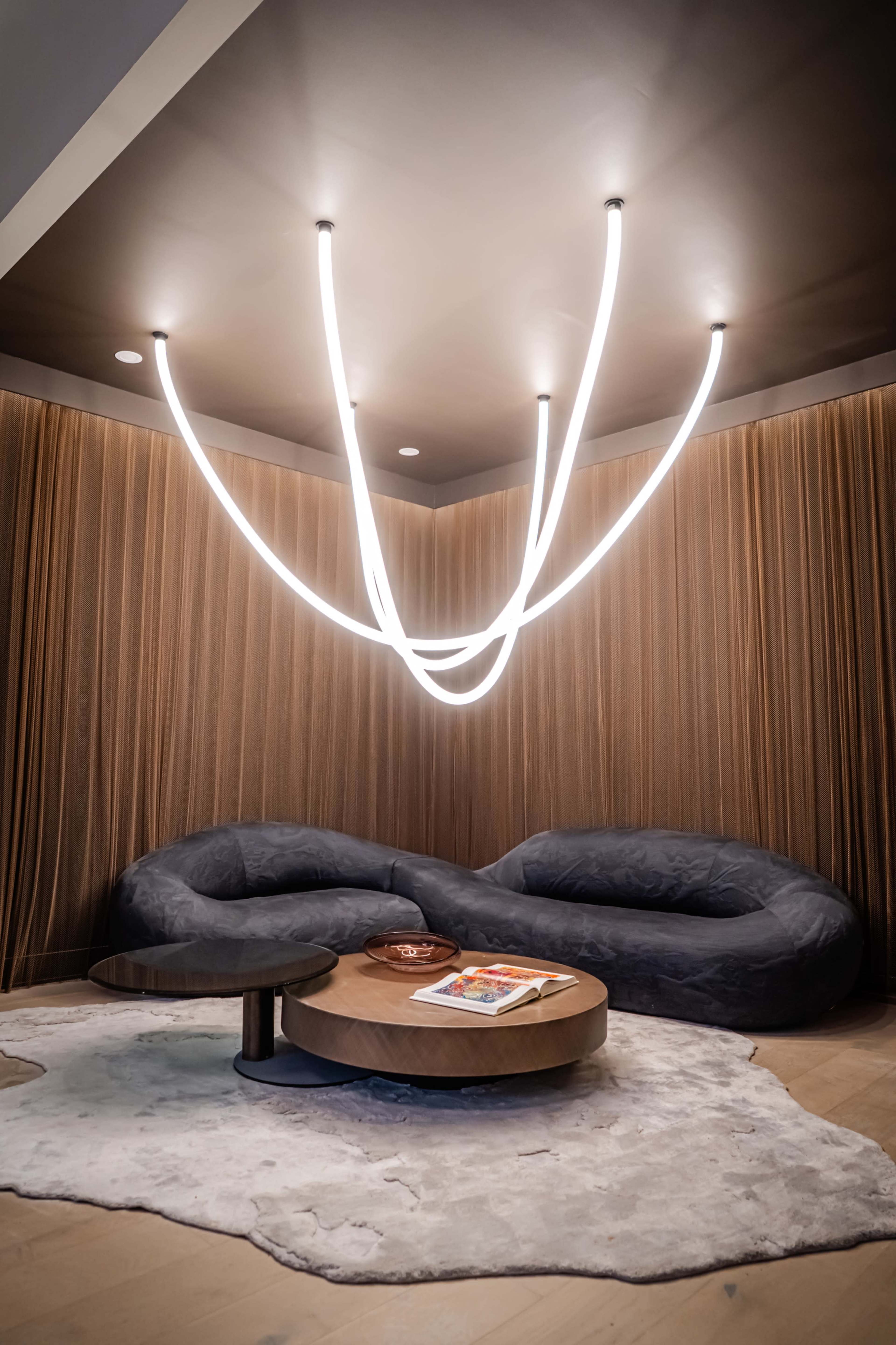 The image shows a modern living space featuring a unique lighting installation overhead, a plush seating arrangement in soft tones, a round coffee table, and a decorative rug on the floor.
