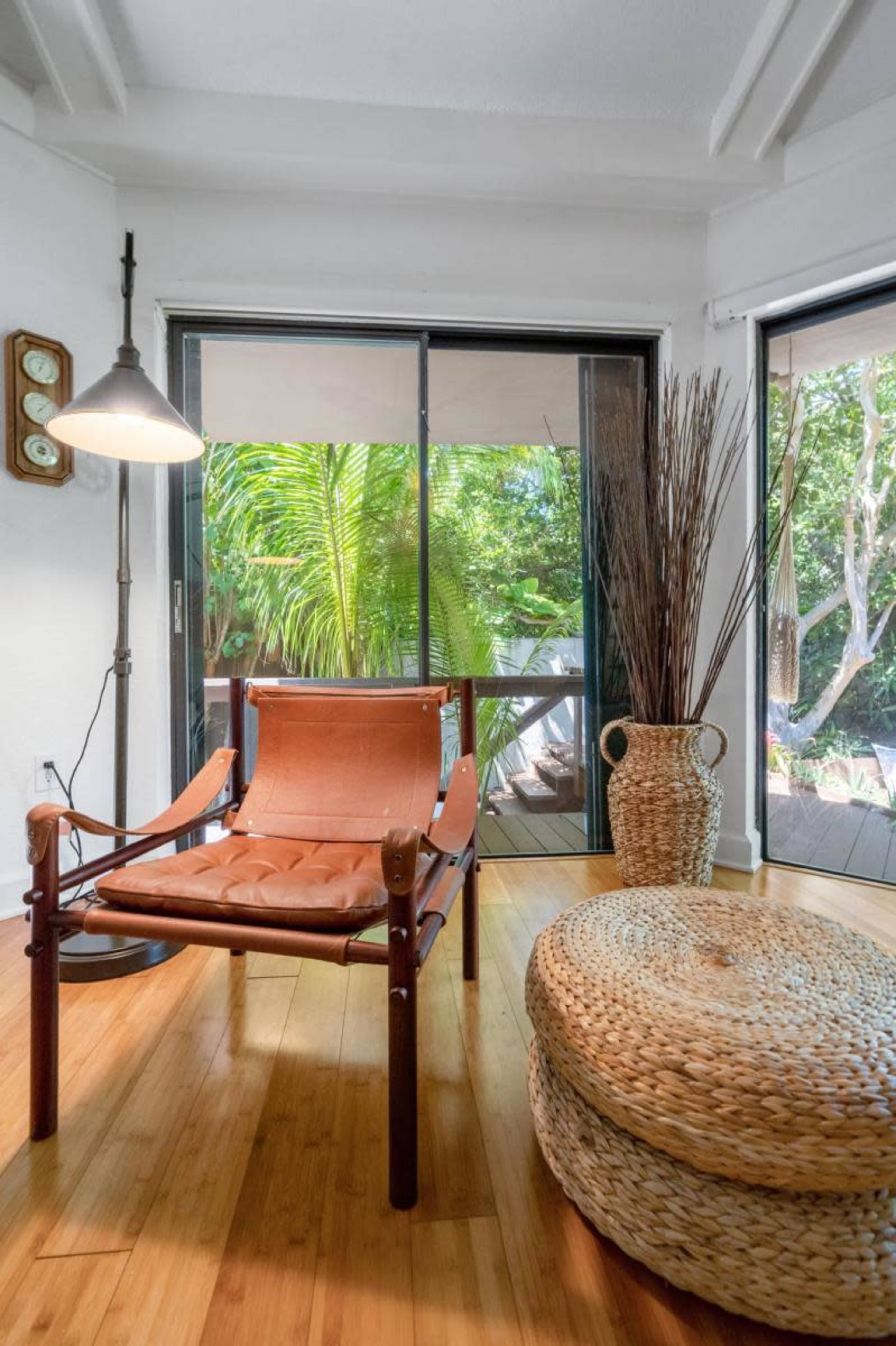 The image shows a modern living space featuring a leather chair next to a floor lamp, a round woven ottoman, and large windows that open to a view of greenery outside.