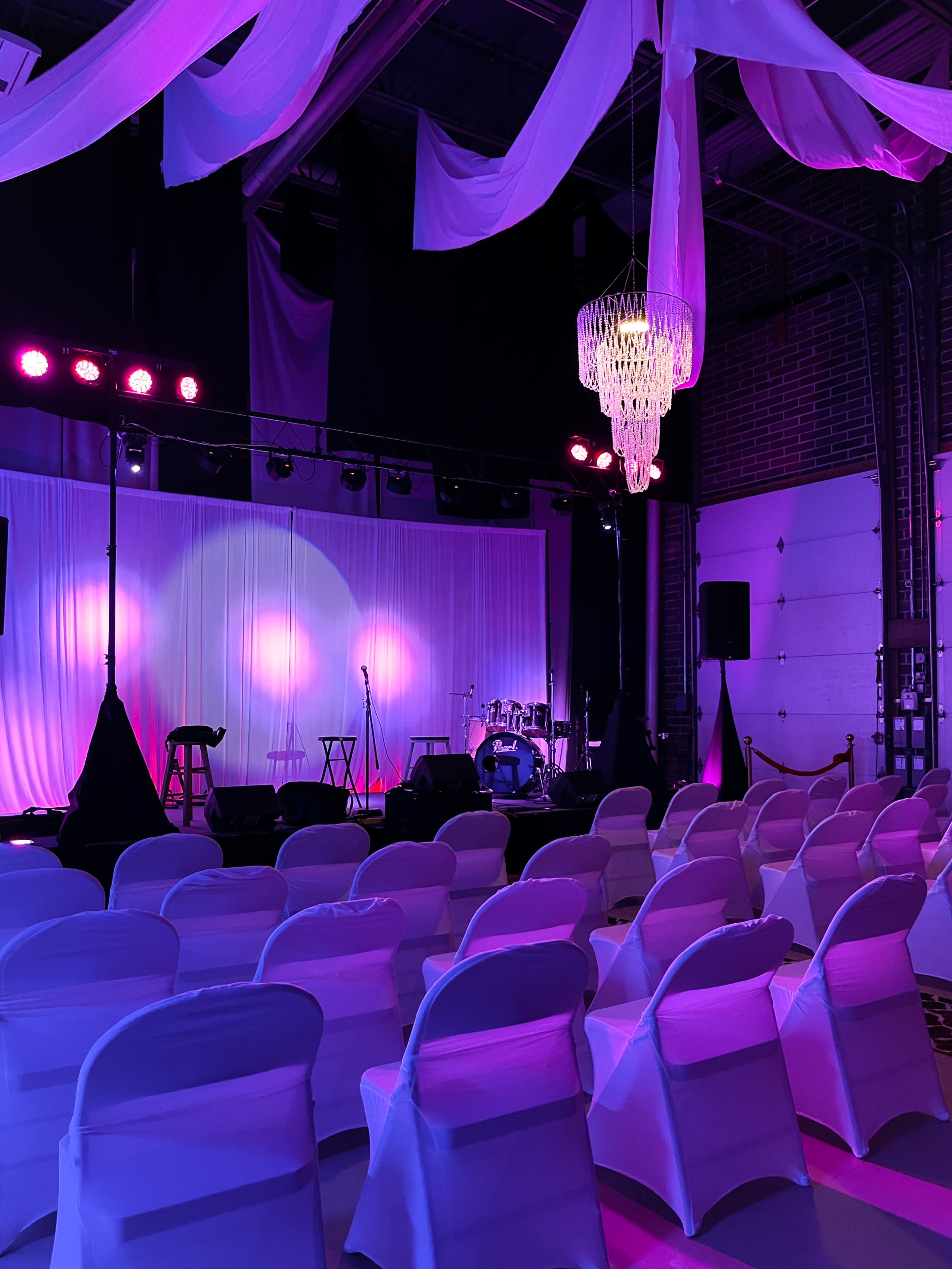 A stage is set up in a dimly lit venue, featuring a drum set and microphones, with rows of white chairs arranged for an audience.