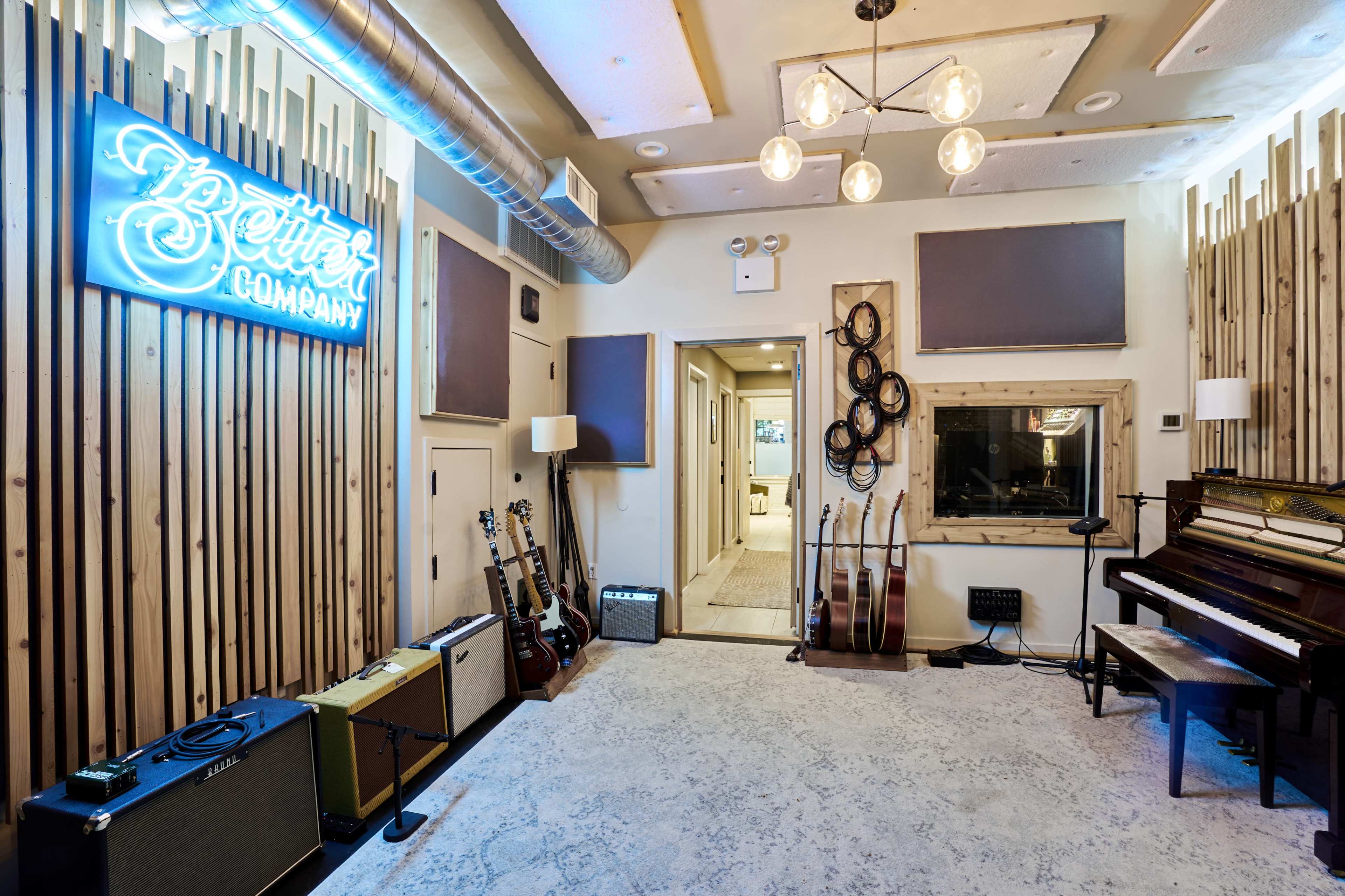 The image shows a music studio with wooden paneling, guitars, amplifiers, a piano, and a neon sign that reads "Better Company."