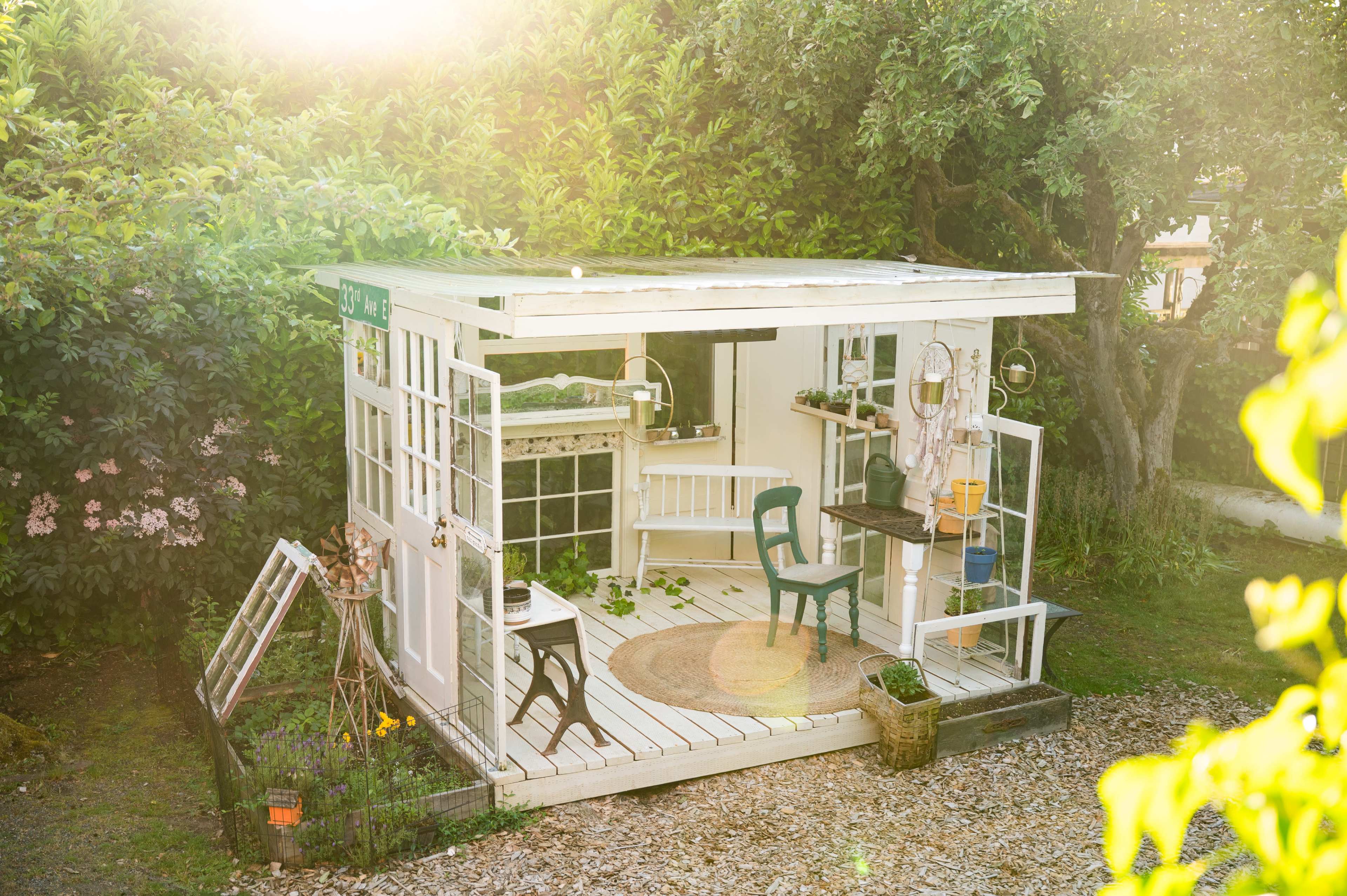 The image shows a small garden shed with large windows, a chair, and various gardening tools arranged outside.