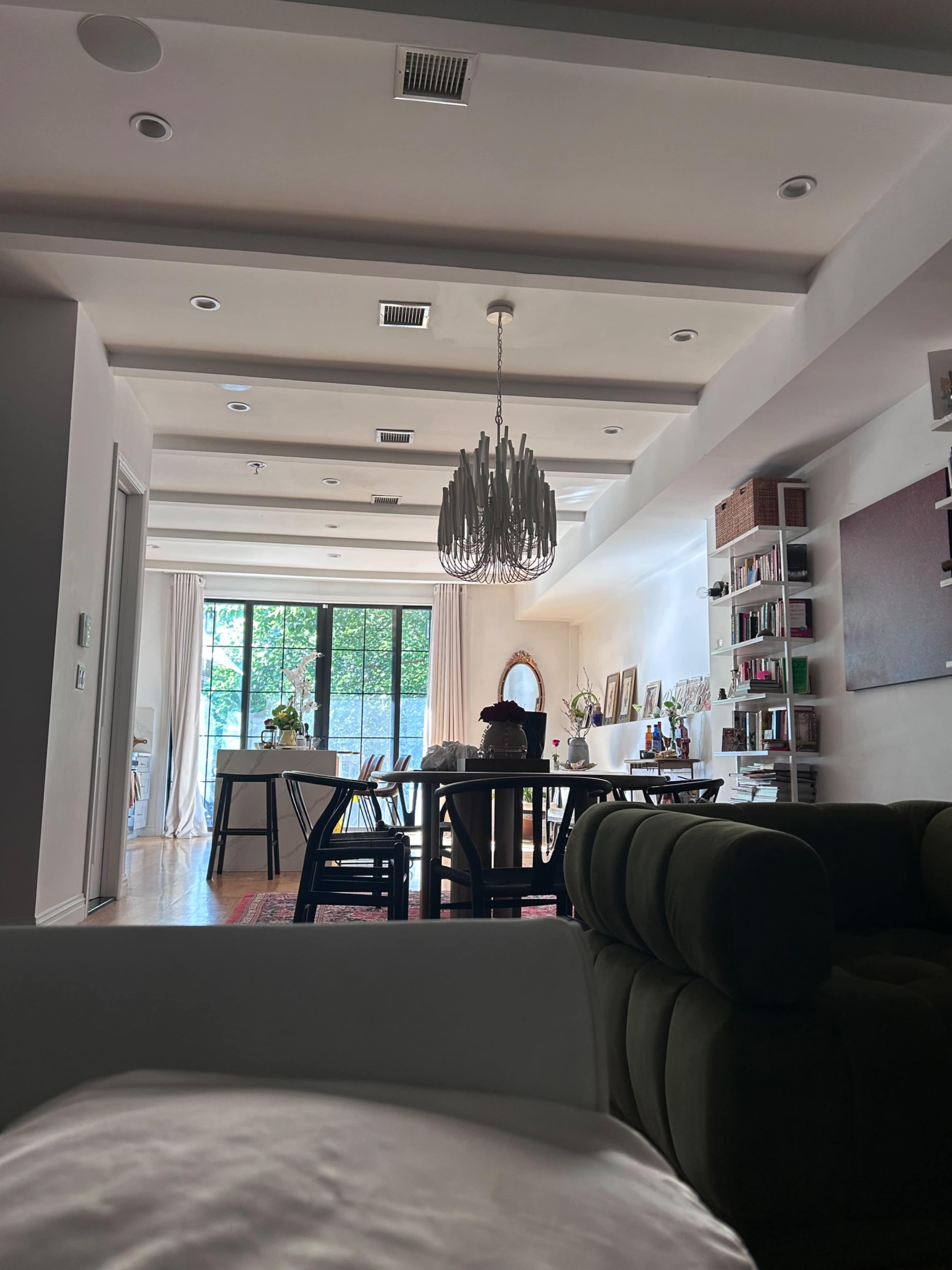 The image shows a spacious living area with a dining table, a chandelier, and large windows leading to a view of greenery outside.