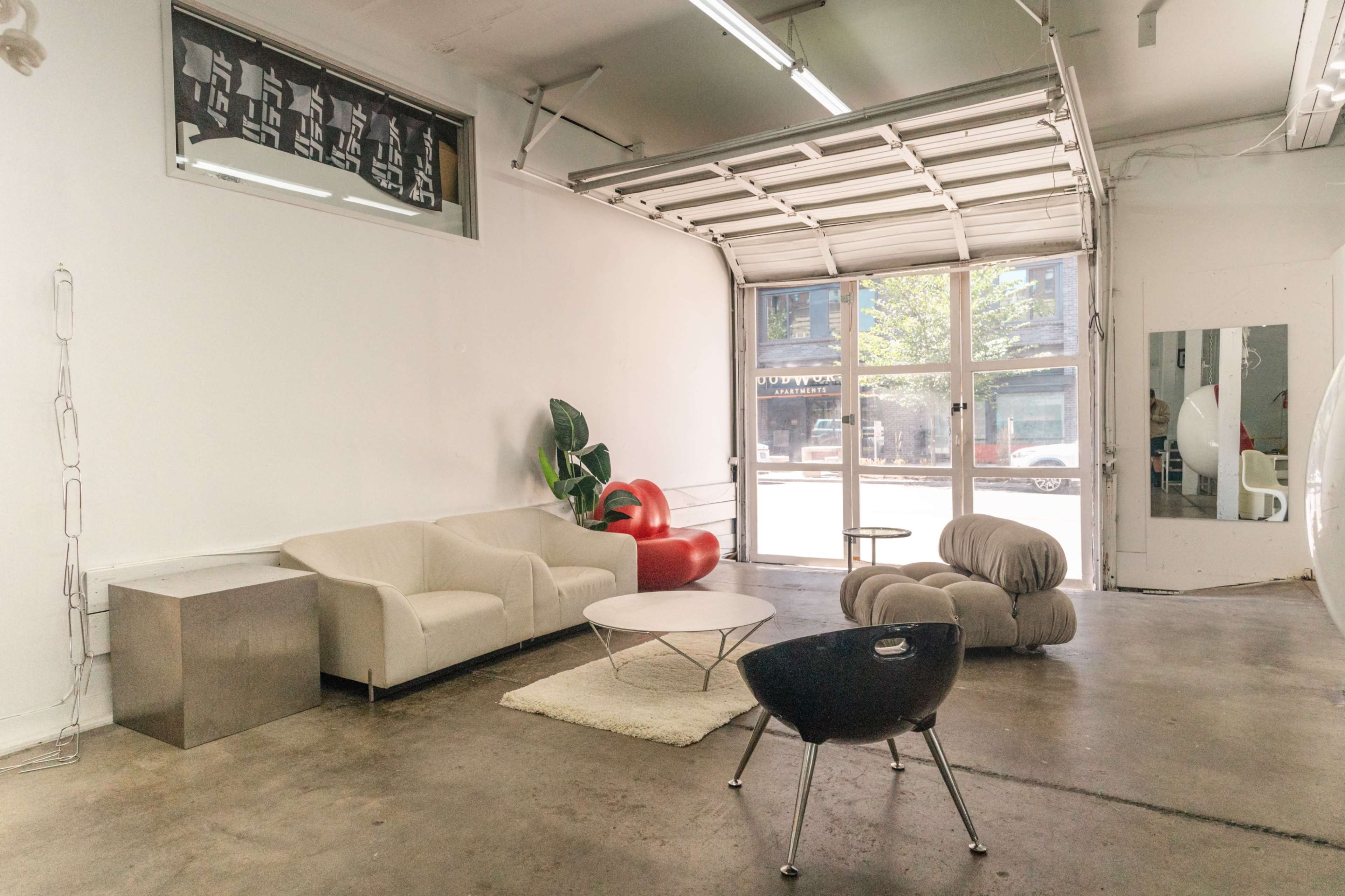 The image shows a minimalist interior of a garage-style space with modern furniture, including a couch, a chair, a coffee table, and large windows letting in natural light.