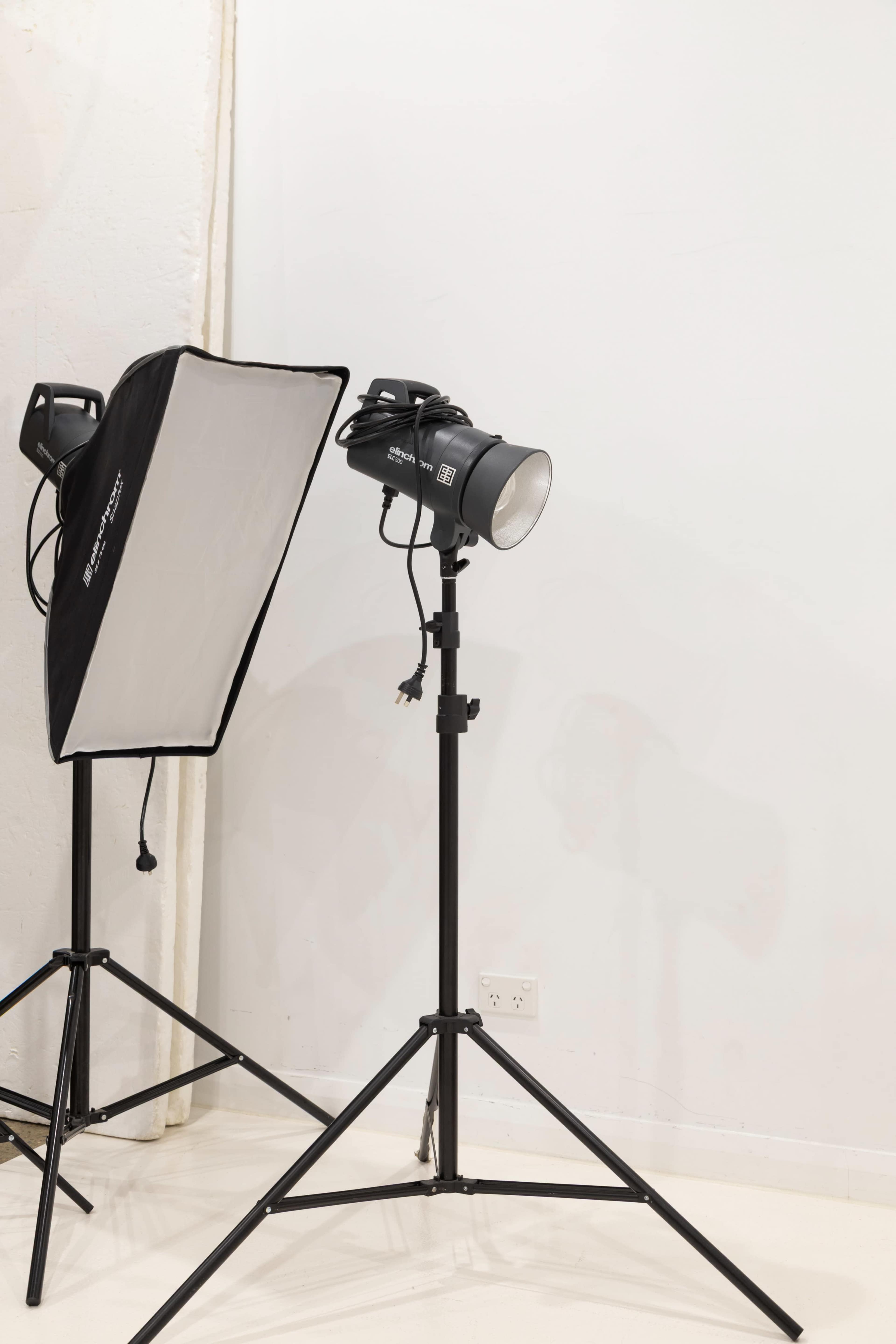 The image shows a studio lighting setup, featuring a light stand with a softbox and another spotlight on a tripod against a plain white wall.