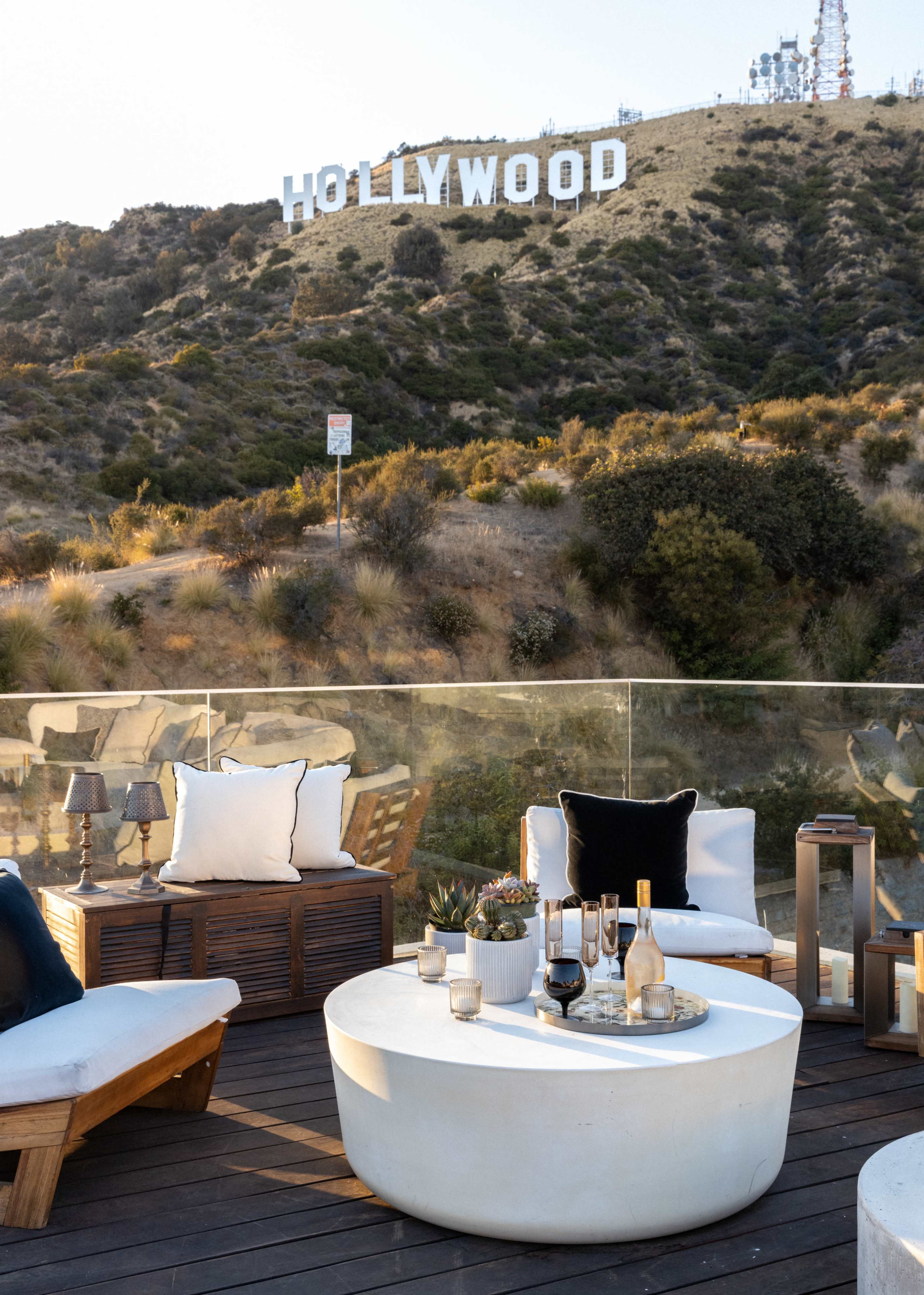 A rooftop seating area with a white circular table and various cushions overlooks the Hollywood sign on a hillside.