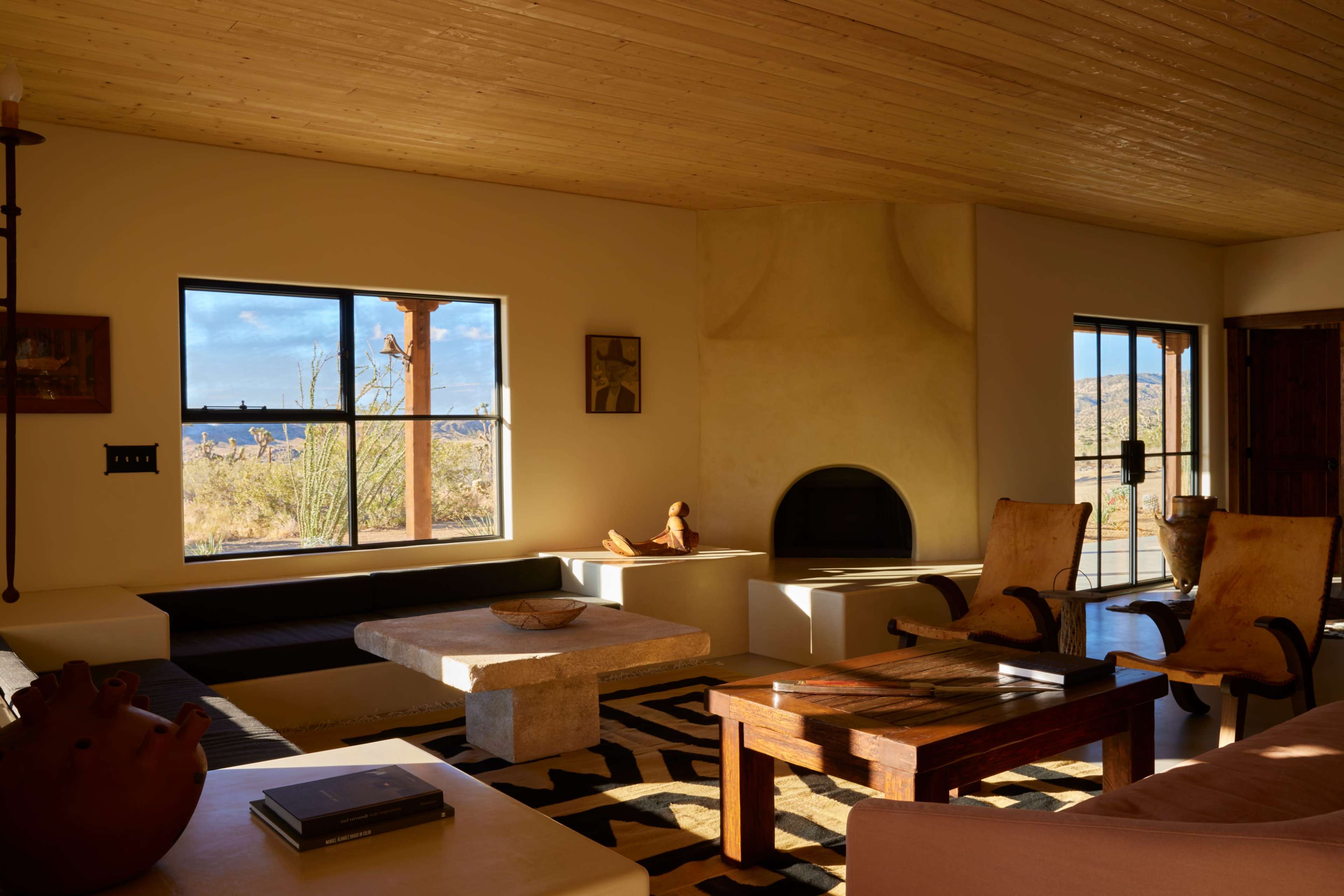 A spacious living room features large windows overlooking a desert landscape, a stone fireplace, and wooden furniture.