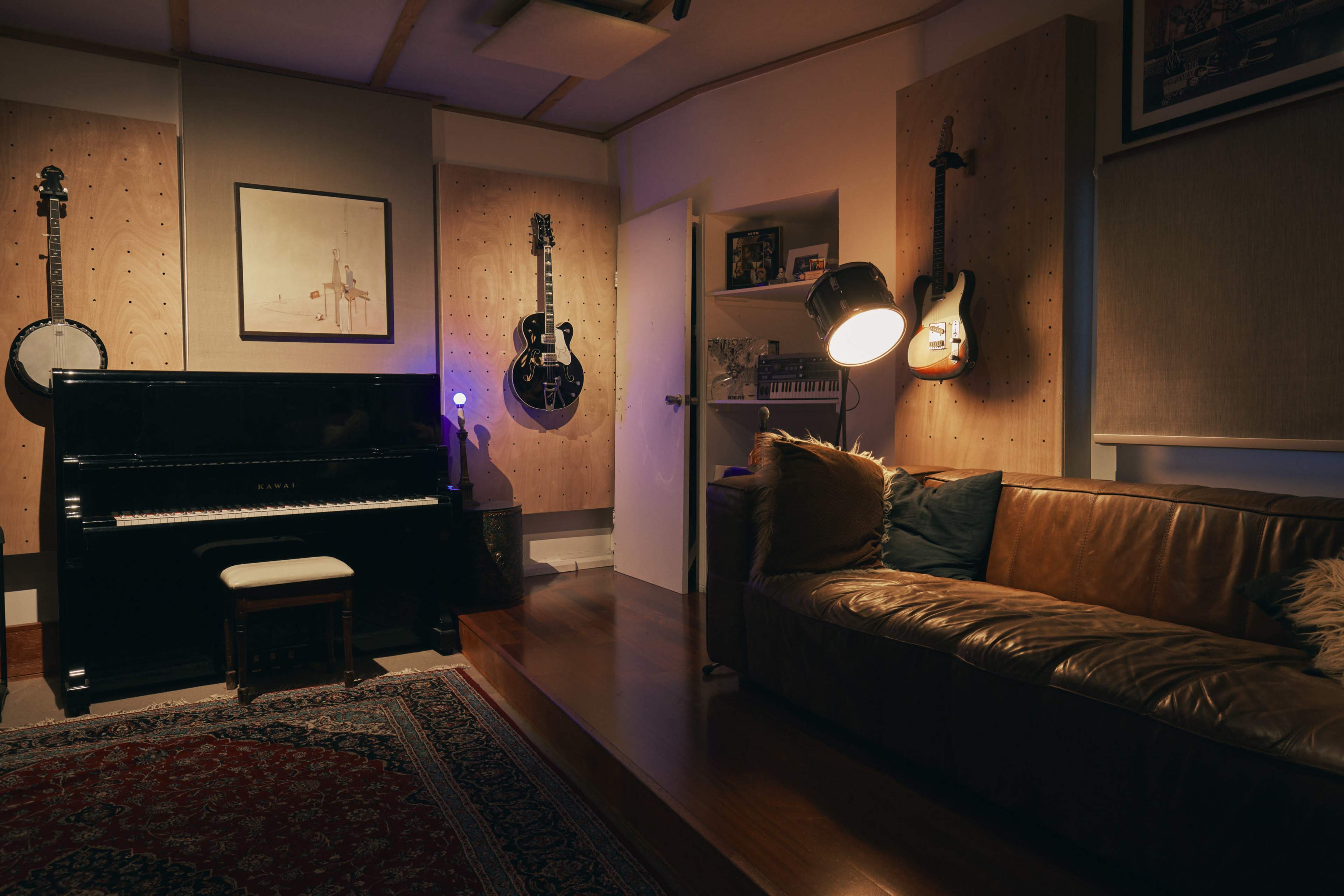 The image features a music studio with a black piano, two guitars mounted on the wall, a brown leather couch, and a floor lamp illuminating the space.