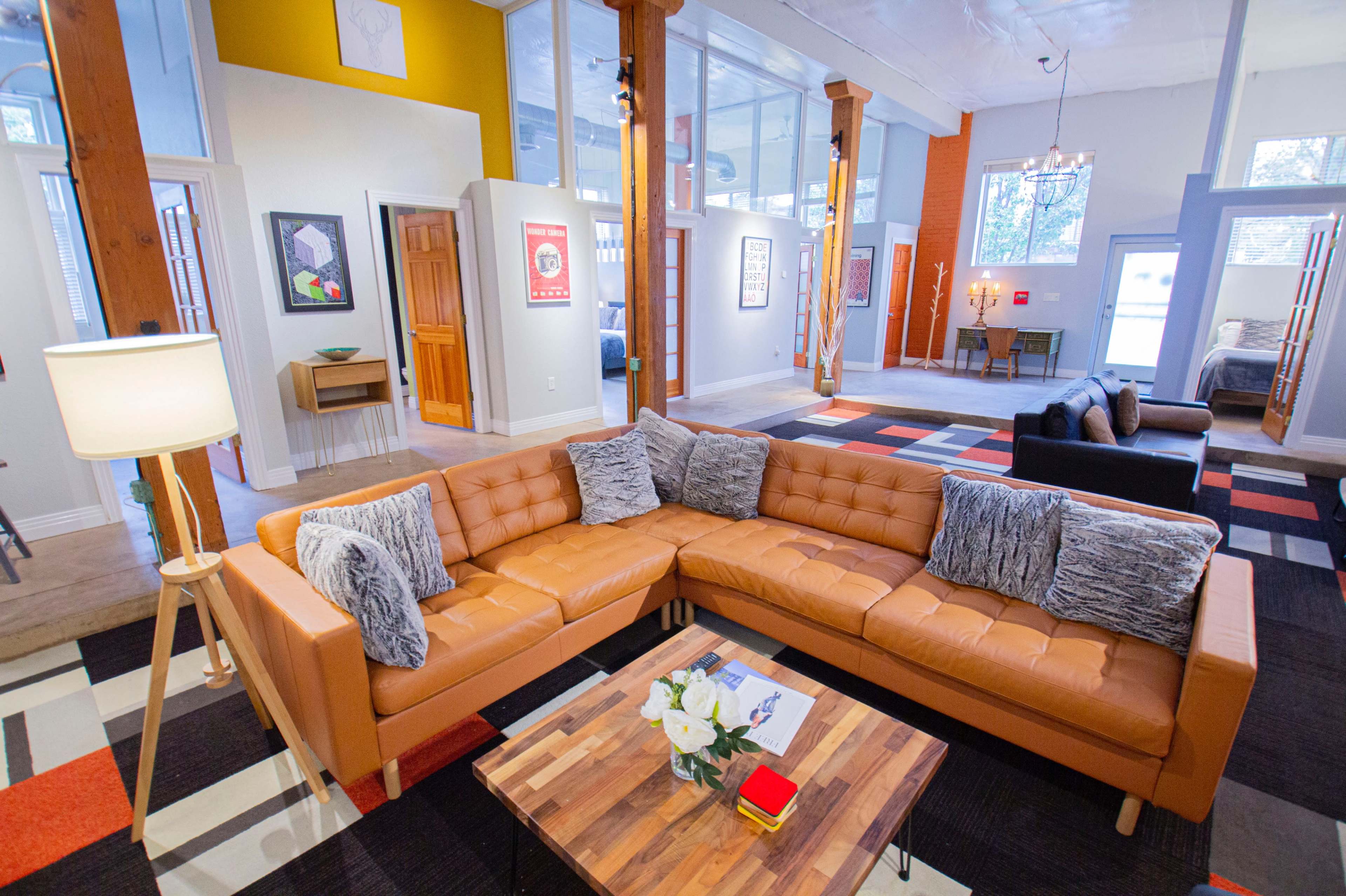 The image depicts a modern living room featuring a brown leather sectional sofa, a wooden coffee table with decorative items, and colorful patterned rugs in a spacious, well-lit area with artwork on the walls.