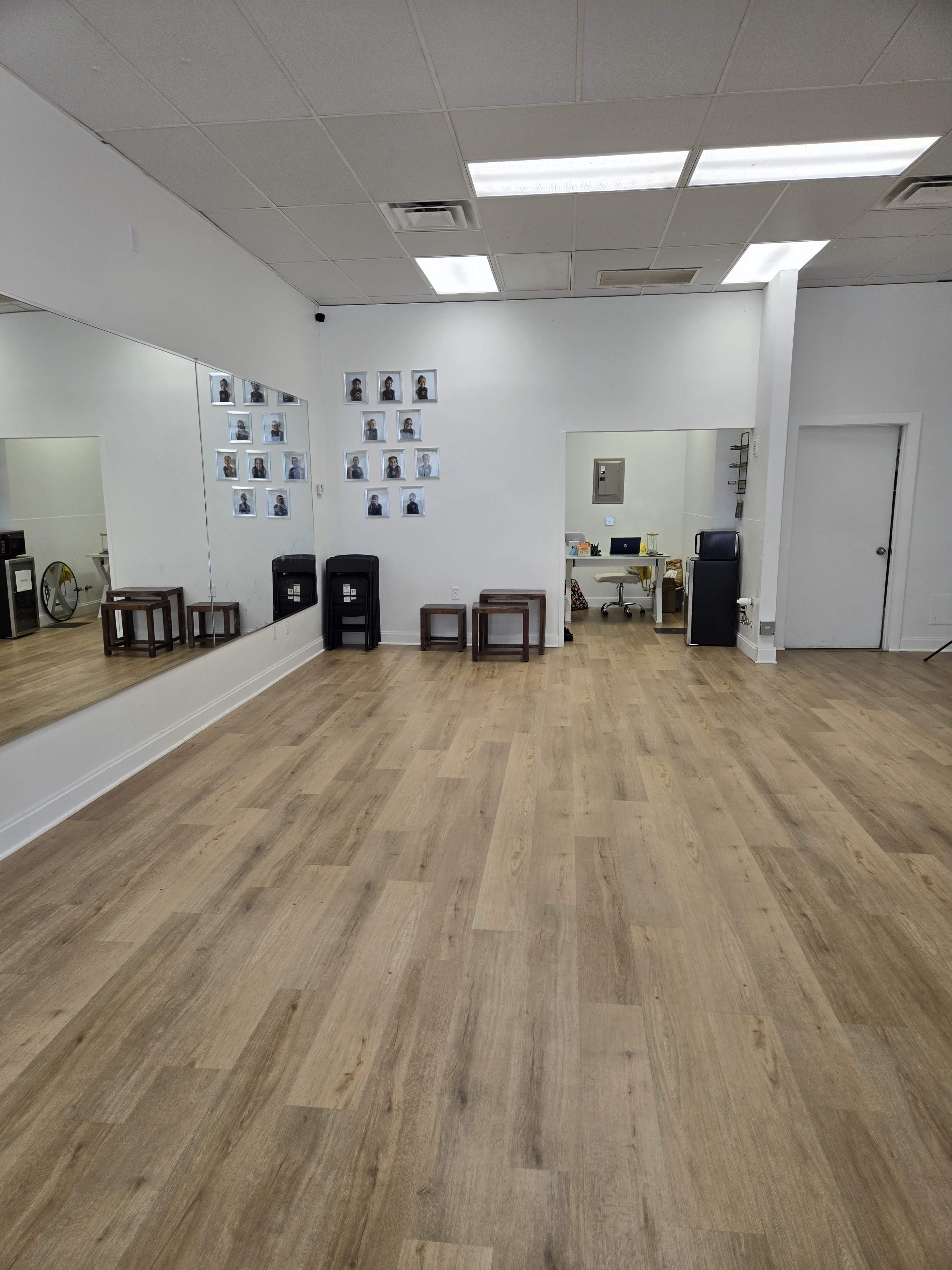 The image shows a spacious room with wooden flooring, two mirrors on one wall, and several framed photos hanging on another wall, along with a small office area visible in the background.