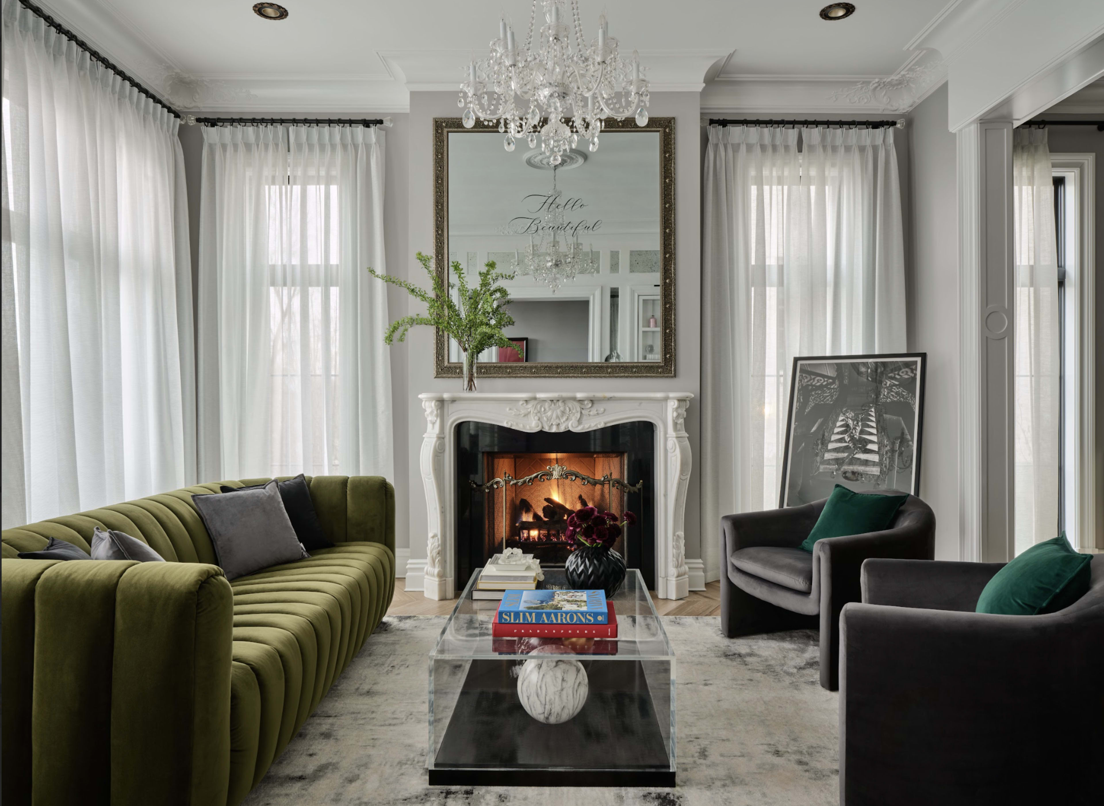 The image features a stylish living room with a green velvet sofa, a modern glass coffee table, two black armchairs, and a decorative fireplace with a large ornate mirror above it.