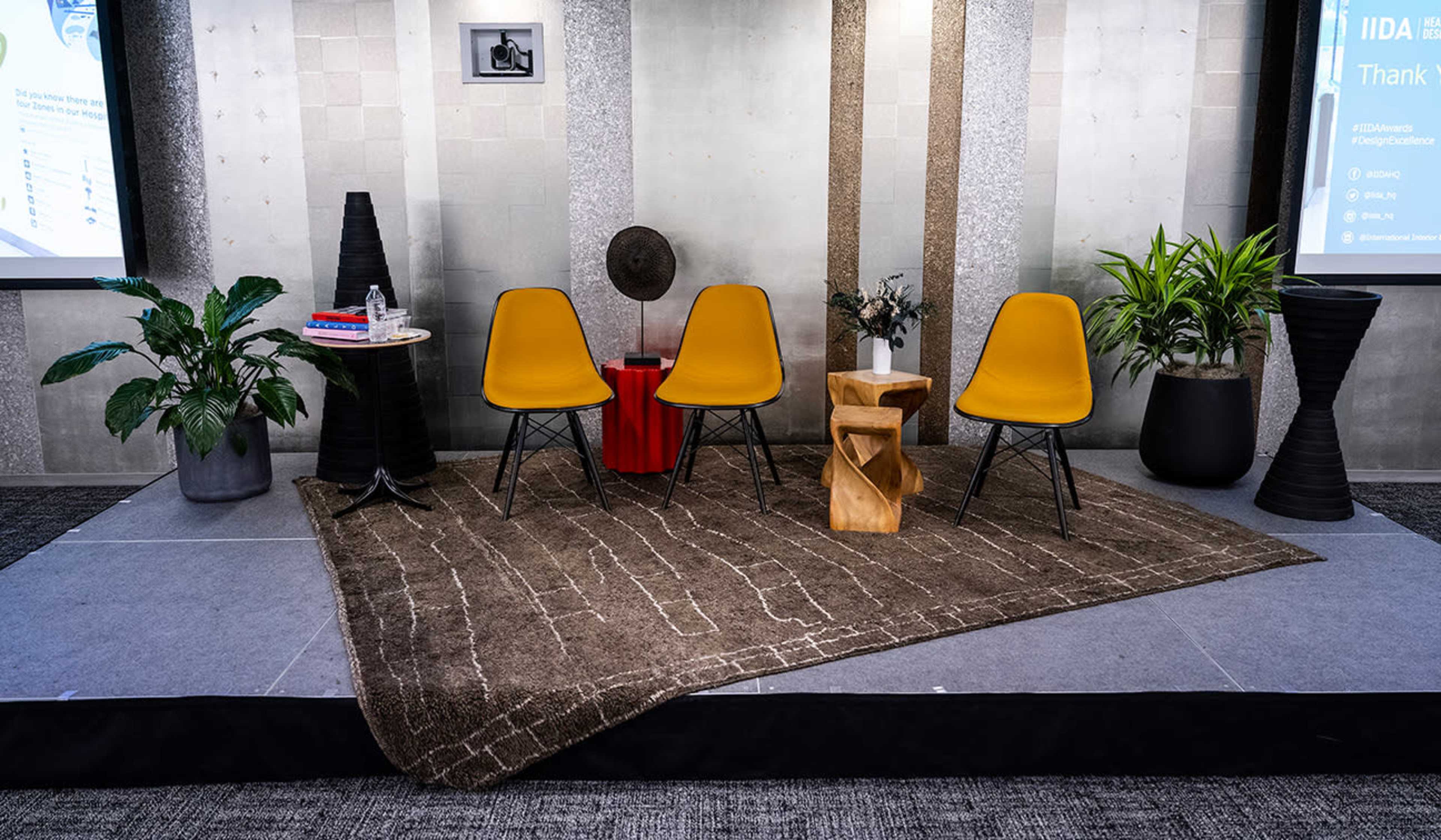 The image shows a minimalist stage setup featuring three yellow chairs, a wooden side table, two planters with plants, and a rug, against a backdrop of grey walls and a screen.