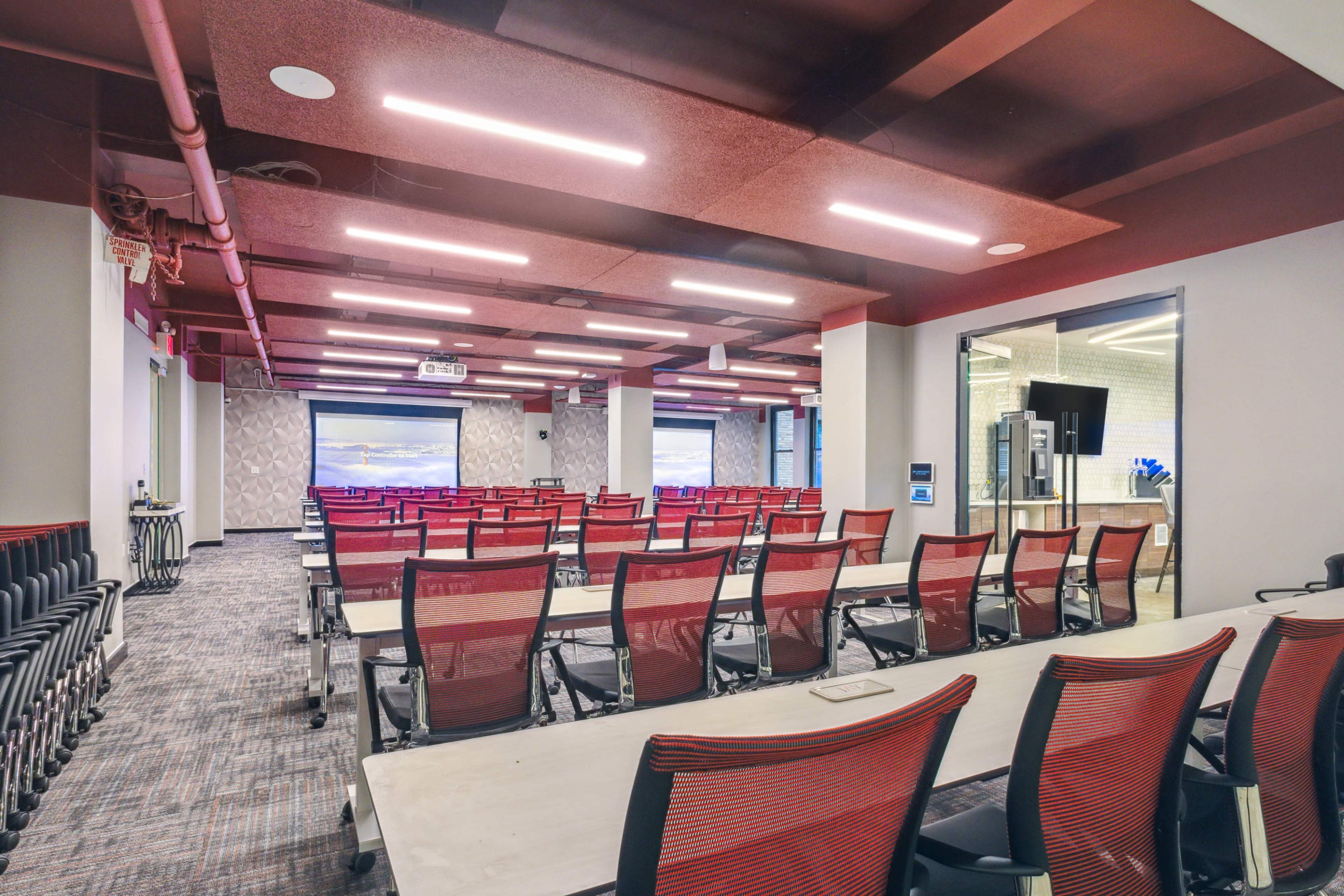A modern conference room with multiple rows of red and white chairs facing a large screen and two smaller screens on the walls.