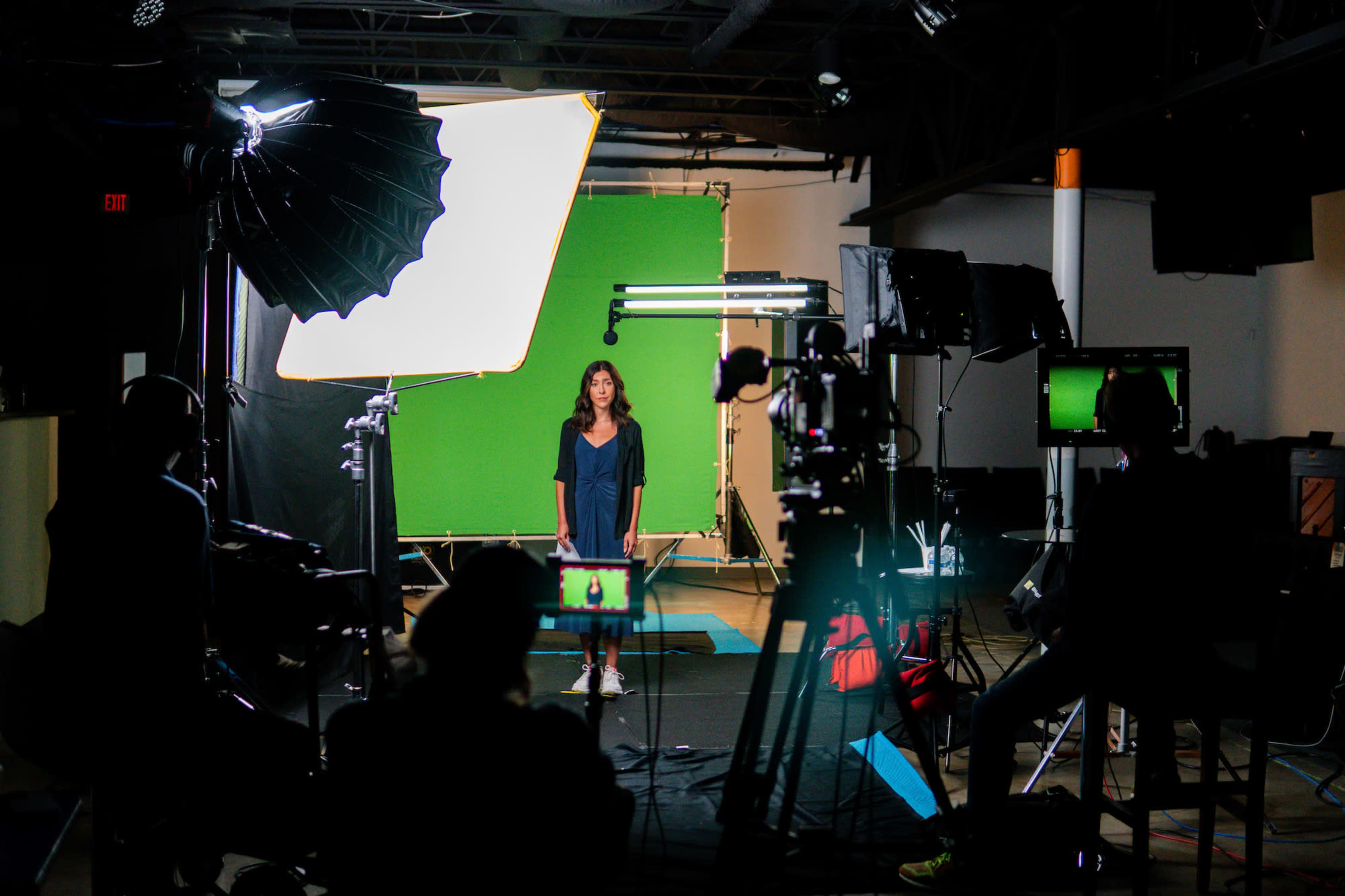 A woman stands in front of a green screen surrounded by studio lighting and camera equipment.