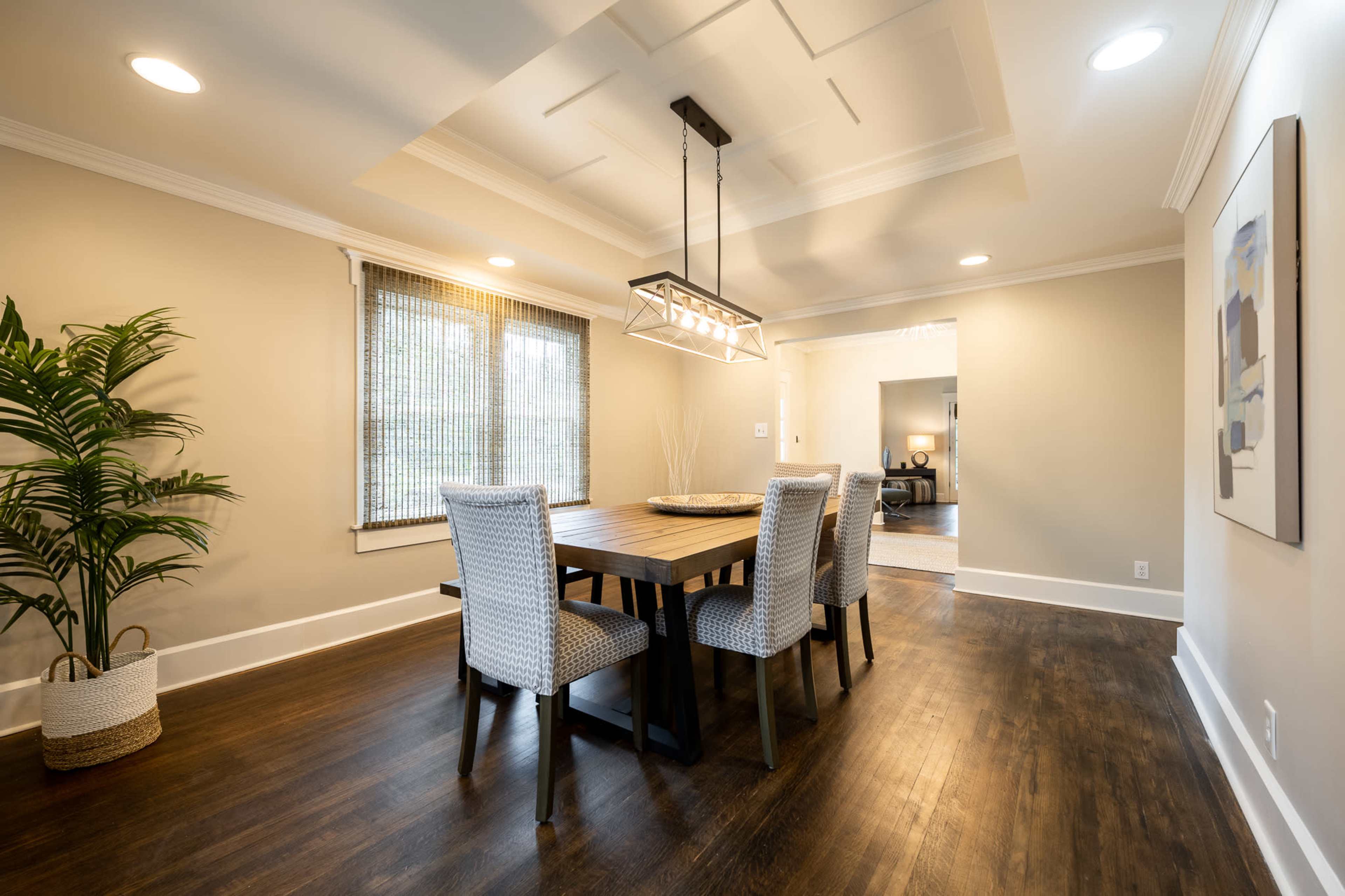 A dining area features a wooden table surrounded by four upholstered chairs, with a modern light fixture hanging above and a potted plant in the corner.