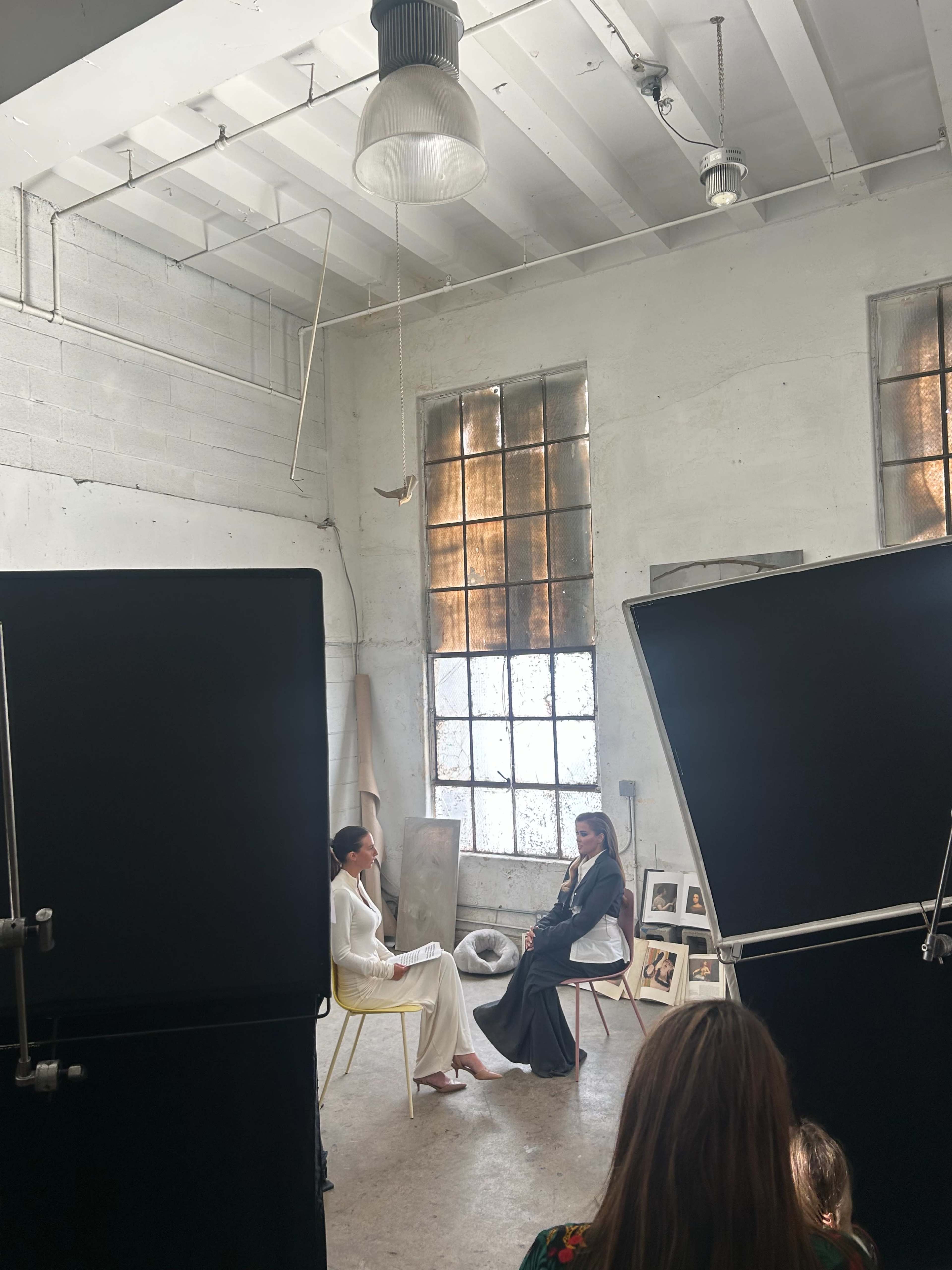 Two women are seated in a well-lit room with large windows, engaged in conversation while a camera crew captures the scene.