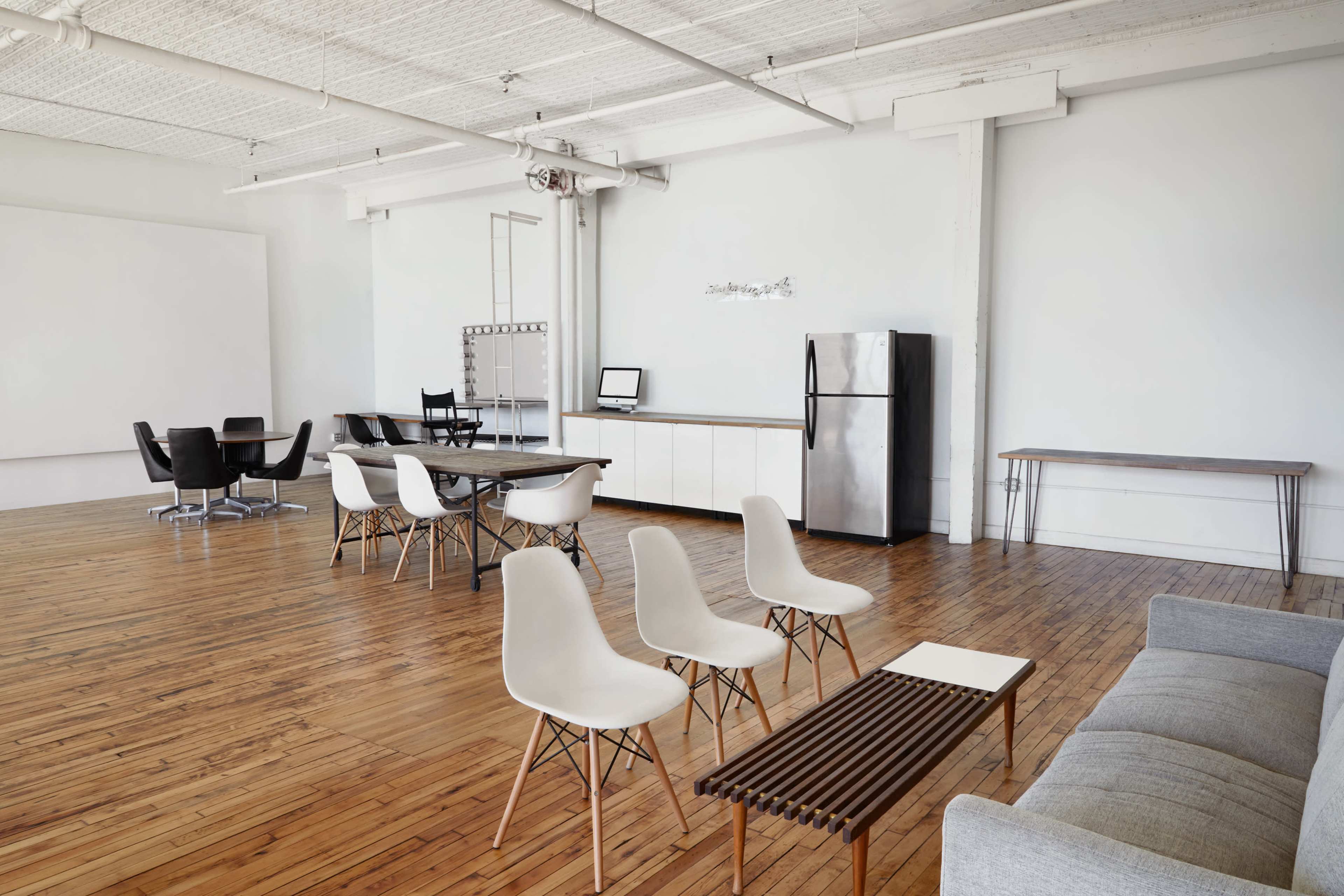 A spacious, minimalist interior with wooden flooring, featuring several chairs, a table, and a kitchenette area with a refrigerator.