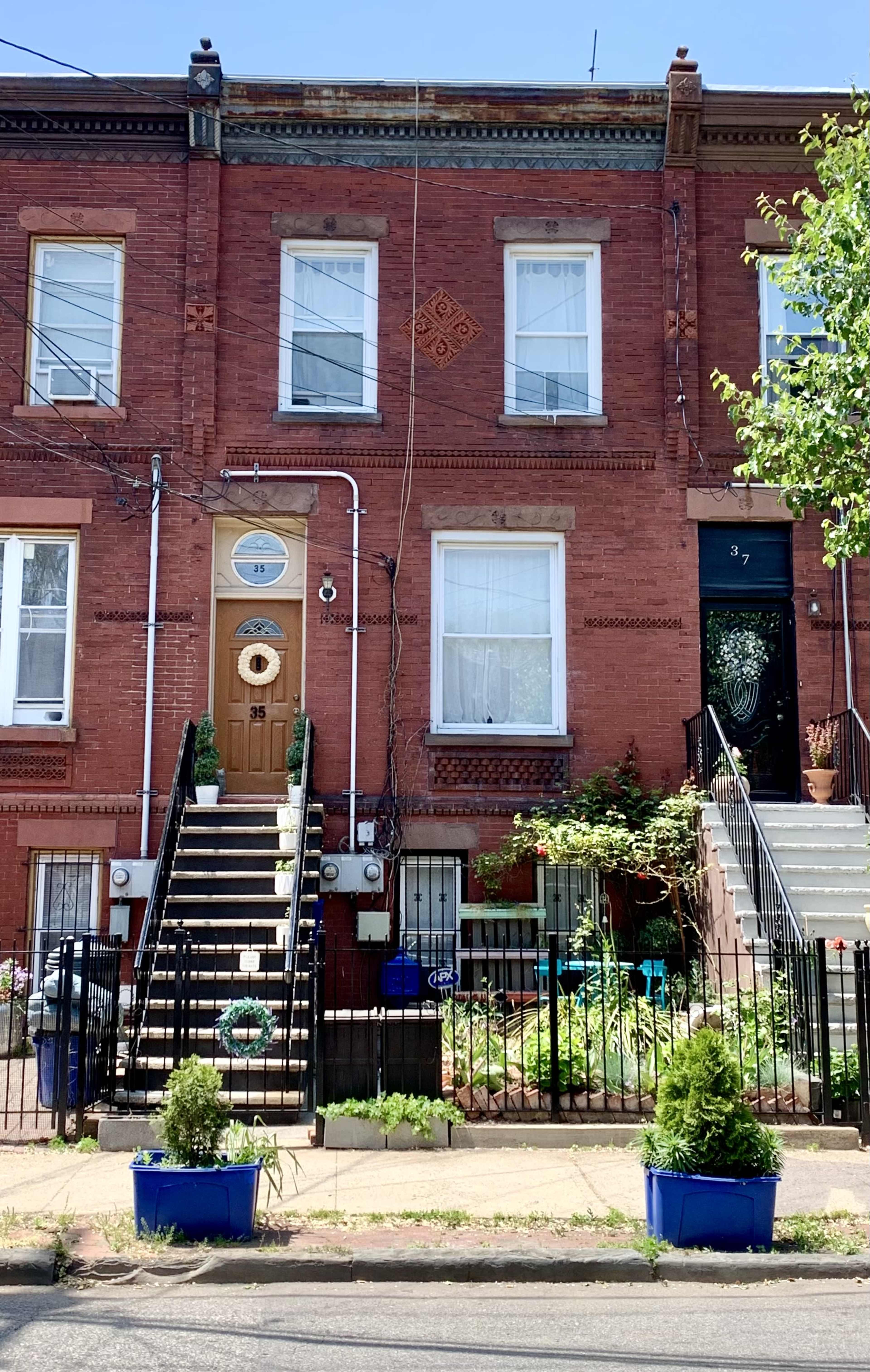 A row of two brick townhouses features distinct front doors and stairs, with greenery and potted plants visible in the yards.