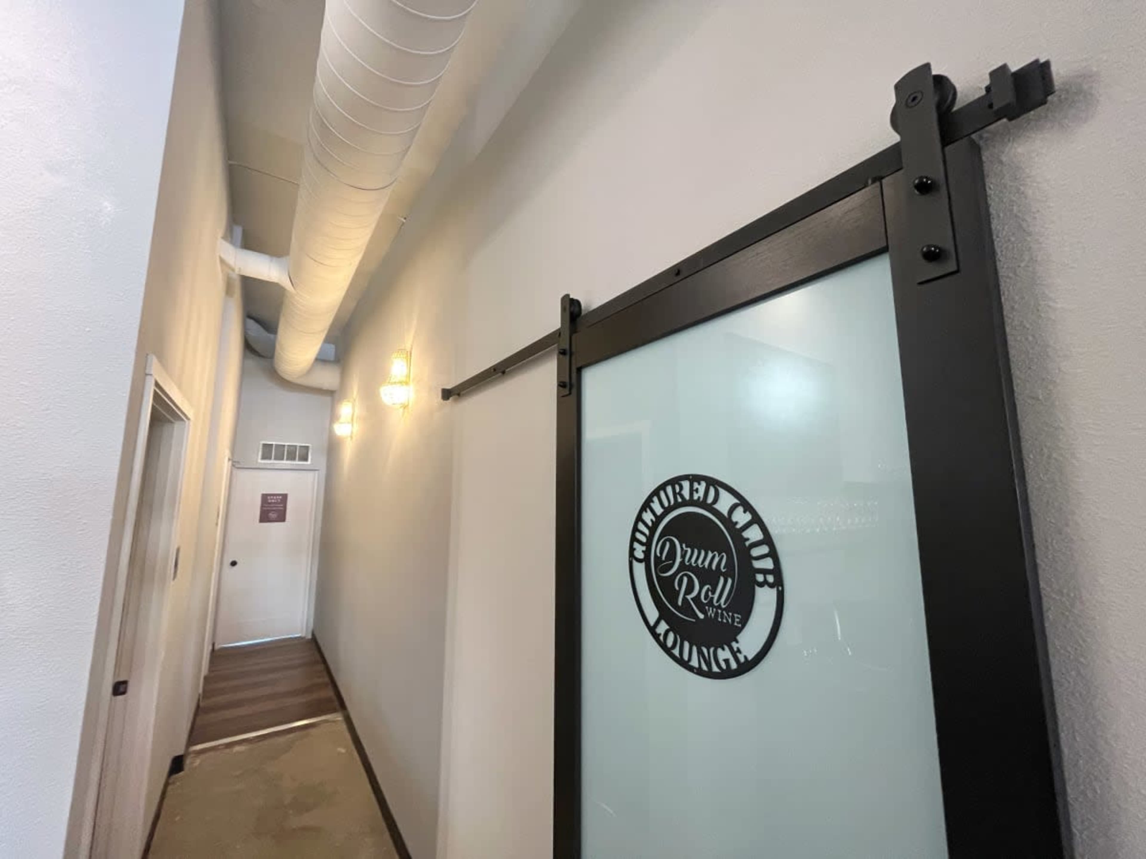 A hallway features a sliding glass door with a circular logo, and exposed pipes along the ceiling.