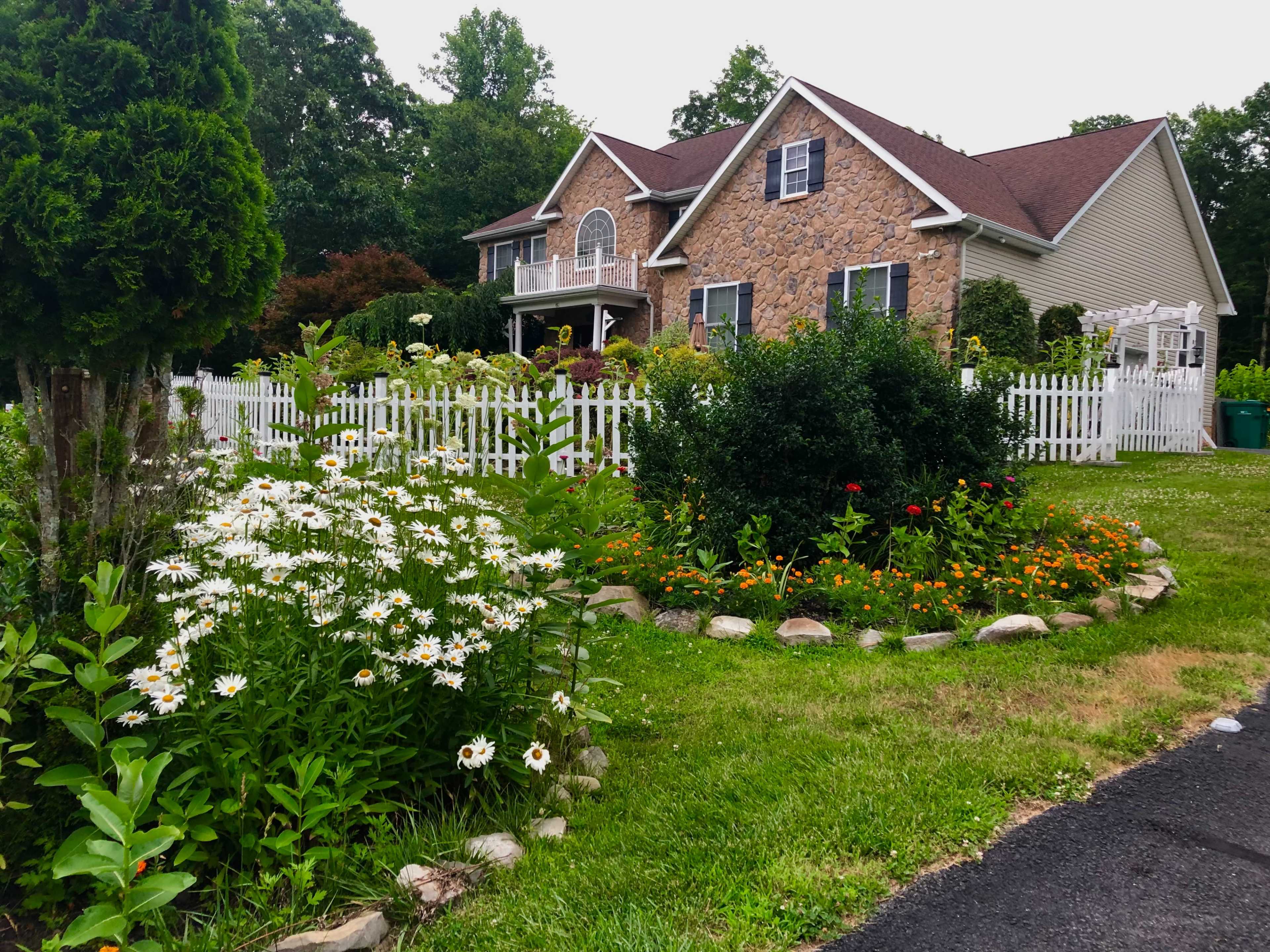 A large two-story house with a stone facade is surrounded by a well-kept garden featuring a circular flower bed and white picket fencing.