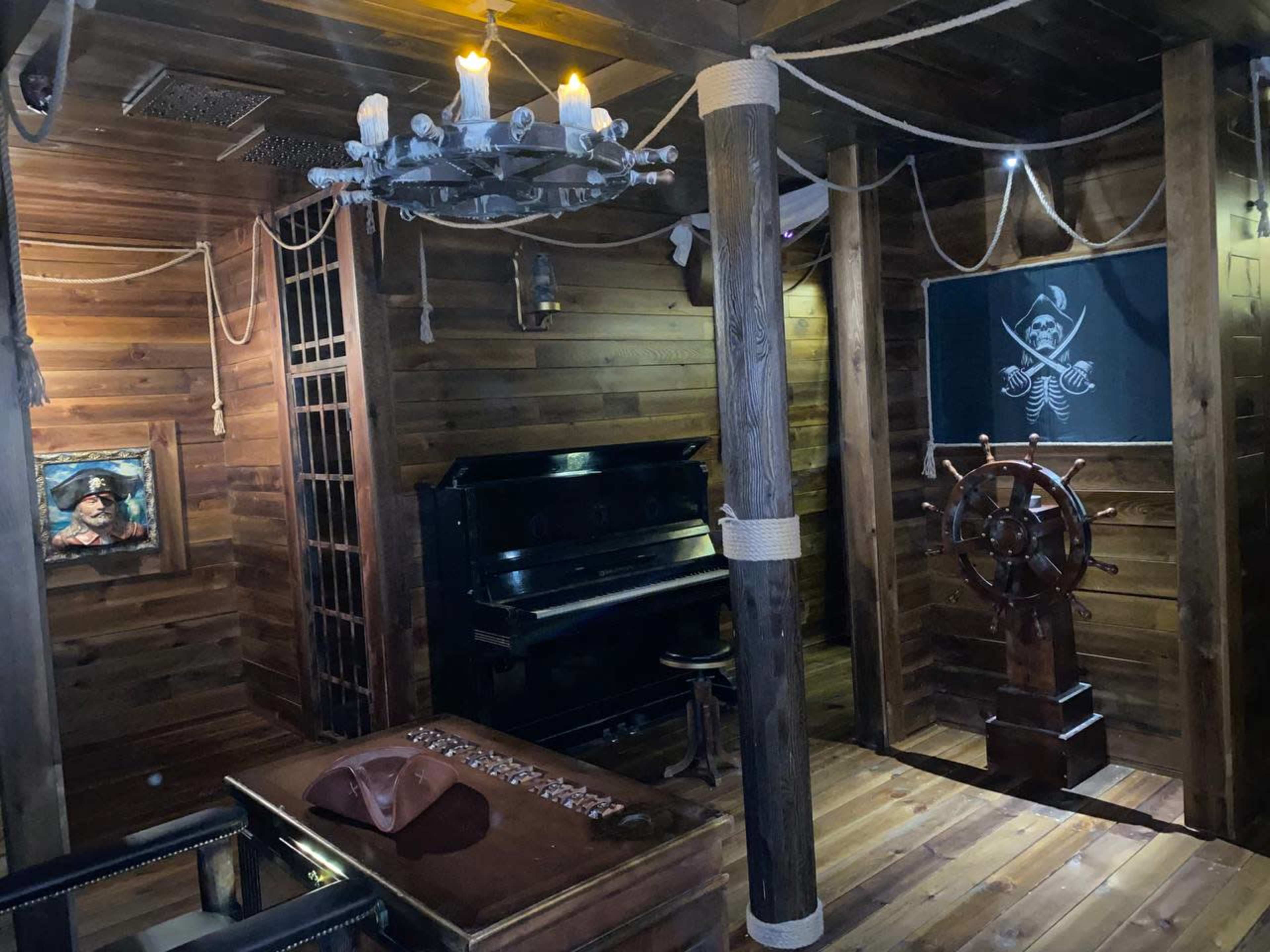 The image shows the interior of a rustic room designed like a pirate ship, featuring a piano, a ship's wheel, and wooden walls with nautical decorations.