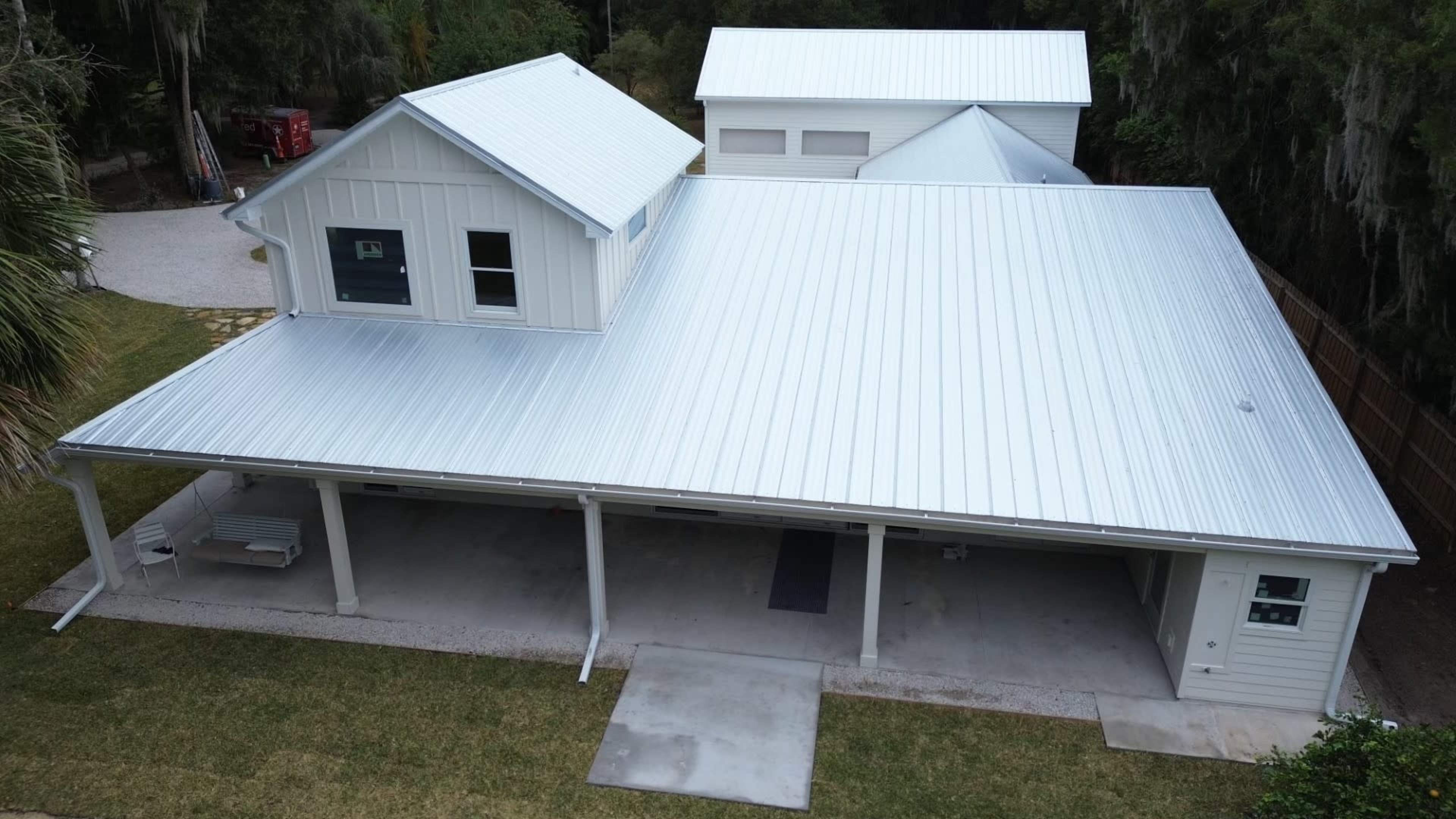 The image shows a modern house with a metal roof, featuring a large covered patio area and surrounded by greenery.