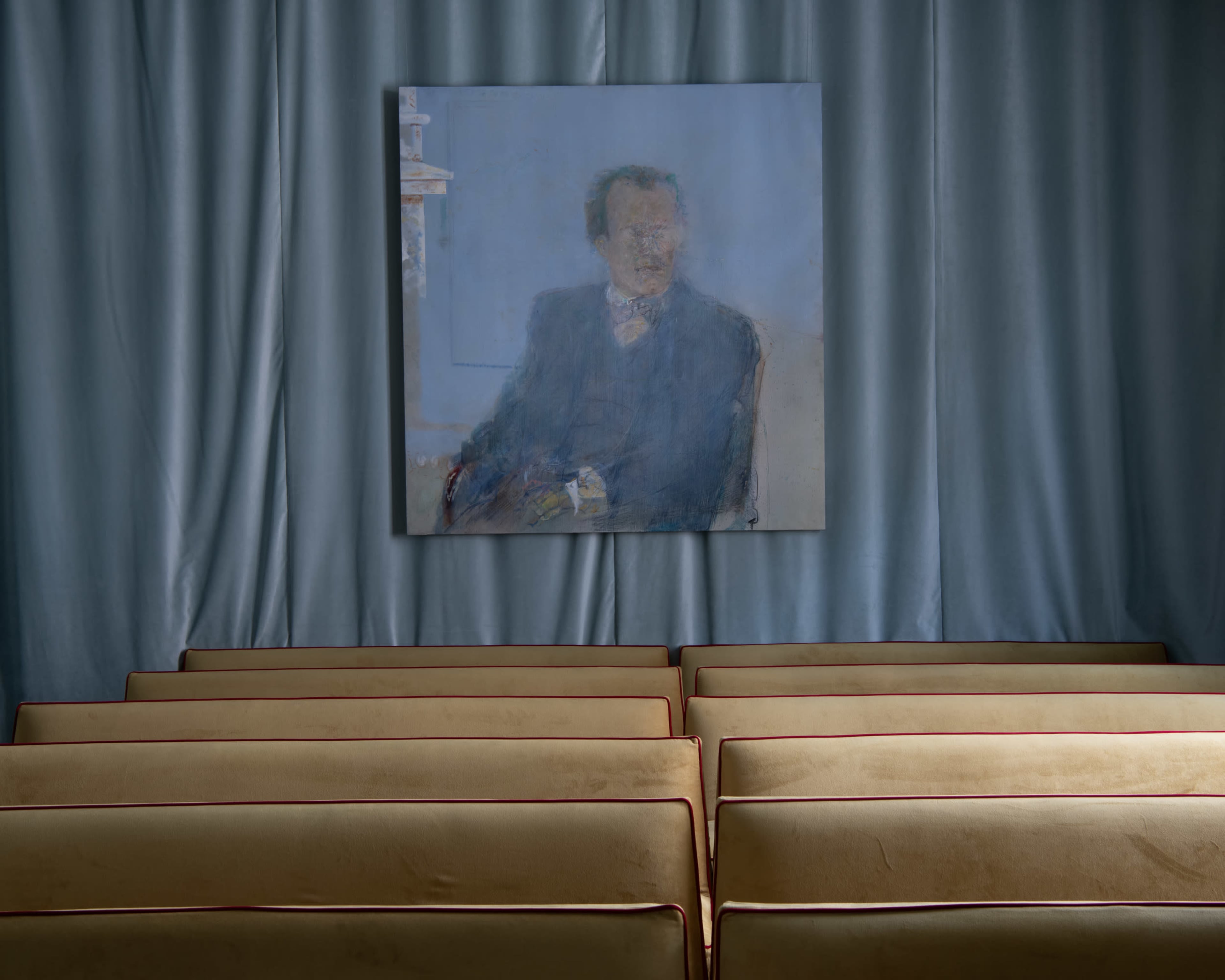 A painting of a seated man hangs on a blue wall, with rows of brown seating visible in the foreground.