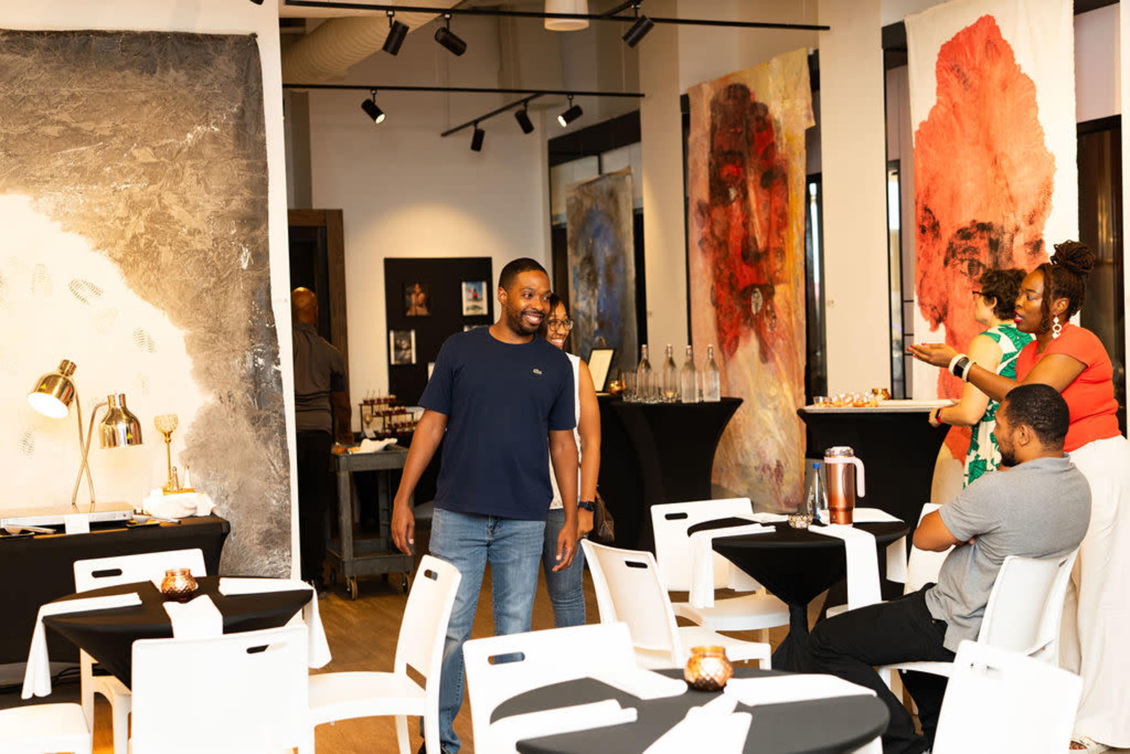 In a contemporary gallery space, individuals interact among black tables adorned with candles, while large abstract artworks hang on the walls.