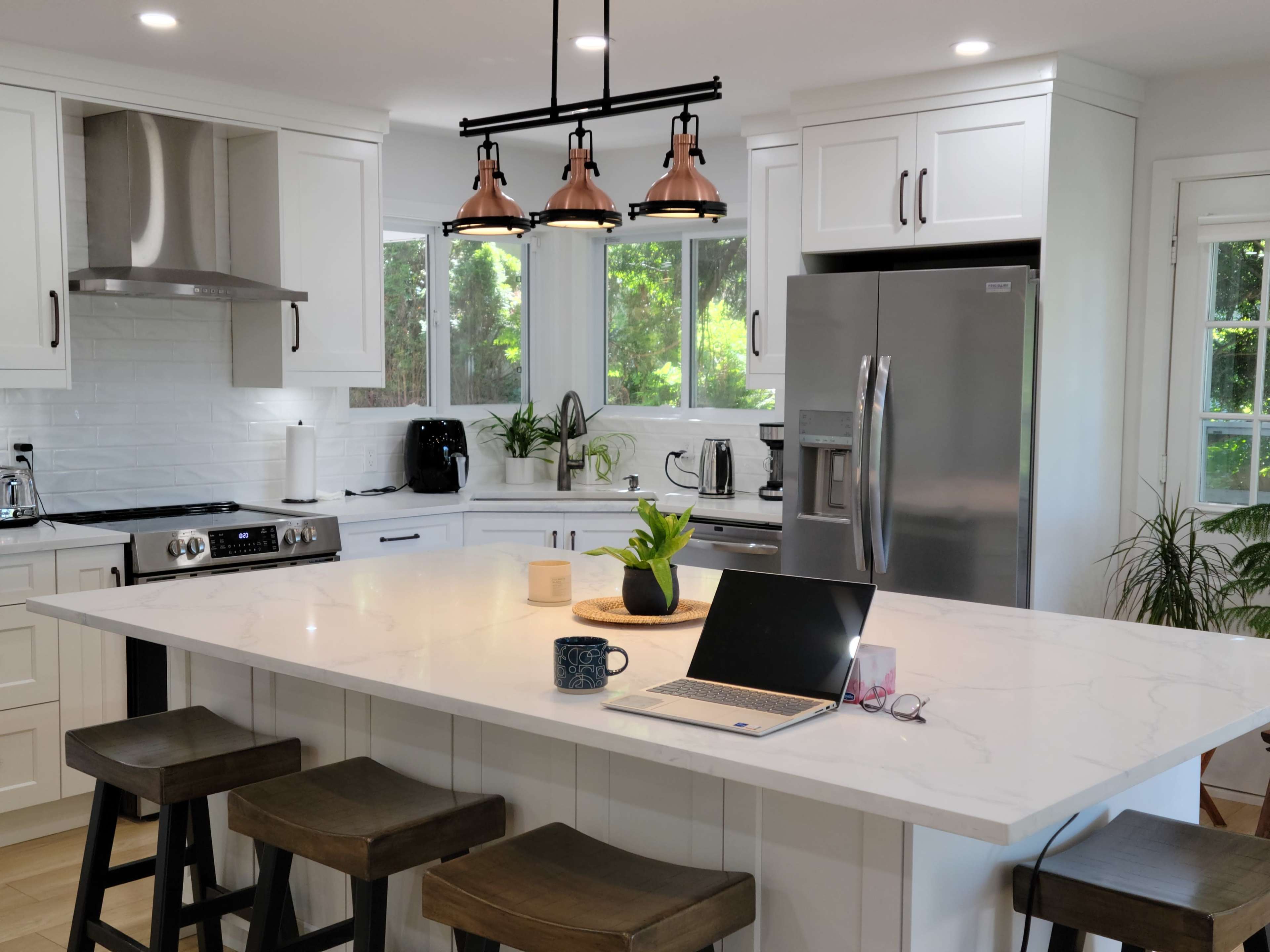 A modern kitchen features white cabinets, a large island with a marble countertop, and a laptop on the surface, surrounded by plants and stainless steel appliances.