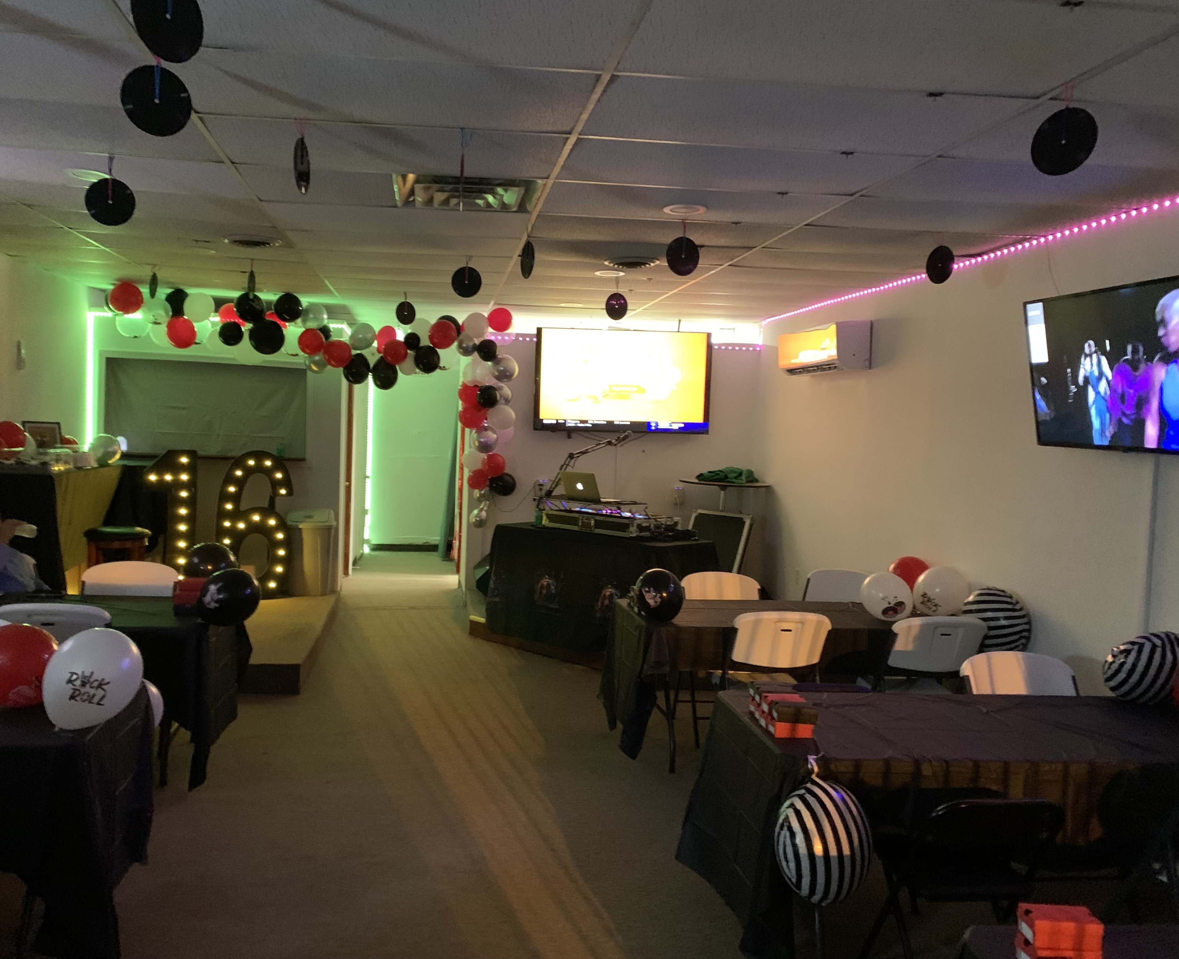 The room is decorated for a party, featuring black, red, and white balloons, a "16" display, and two televisions broadcasting entertainment.