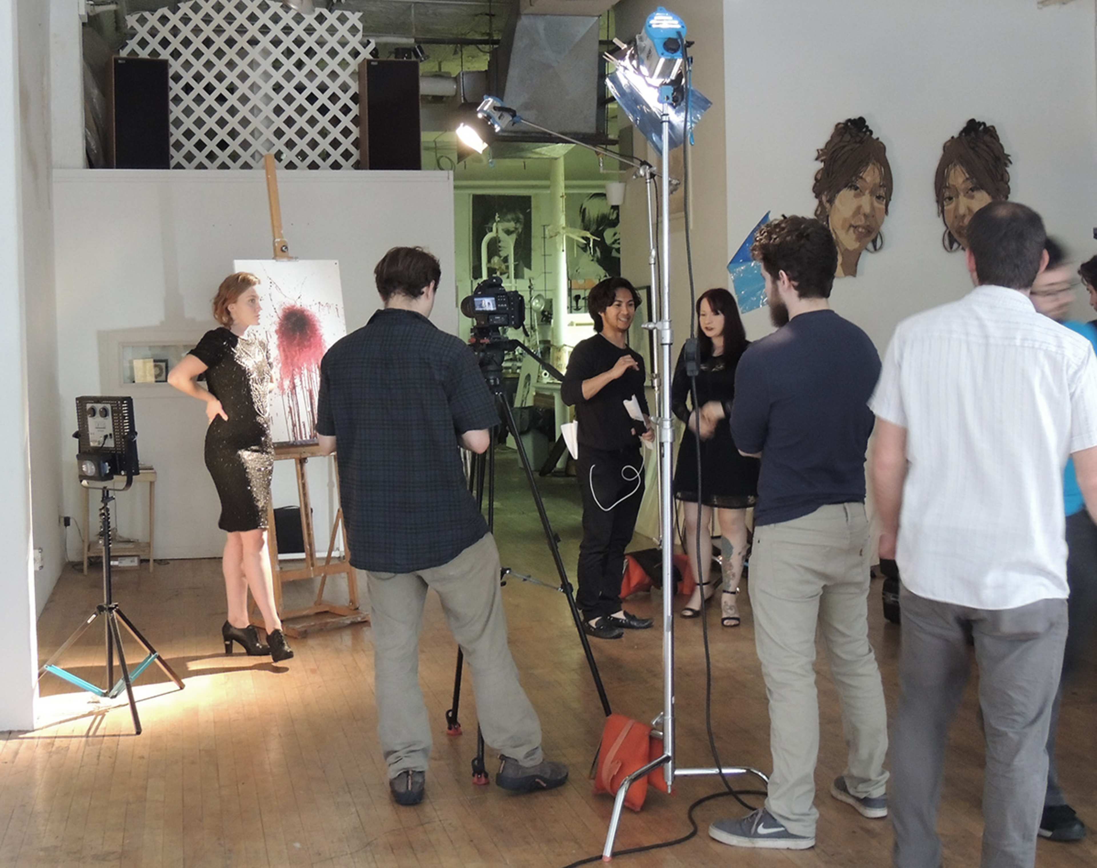 A group of people gathers in an art studio while a camera crew films a woman posing in front of an easel with a painted backdrop.