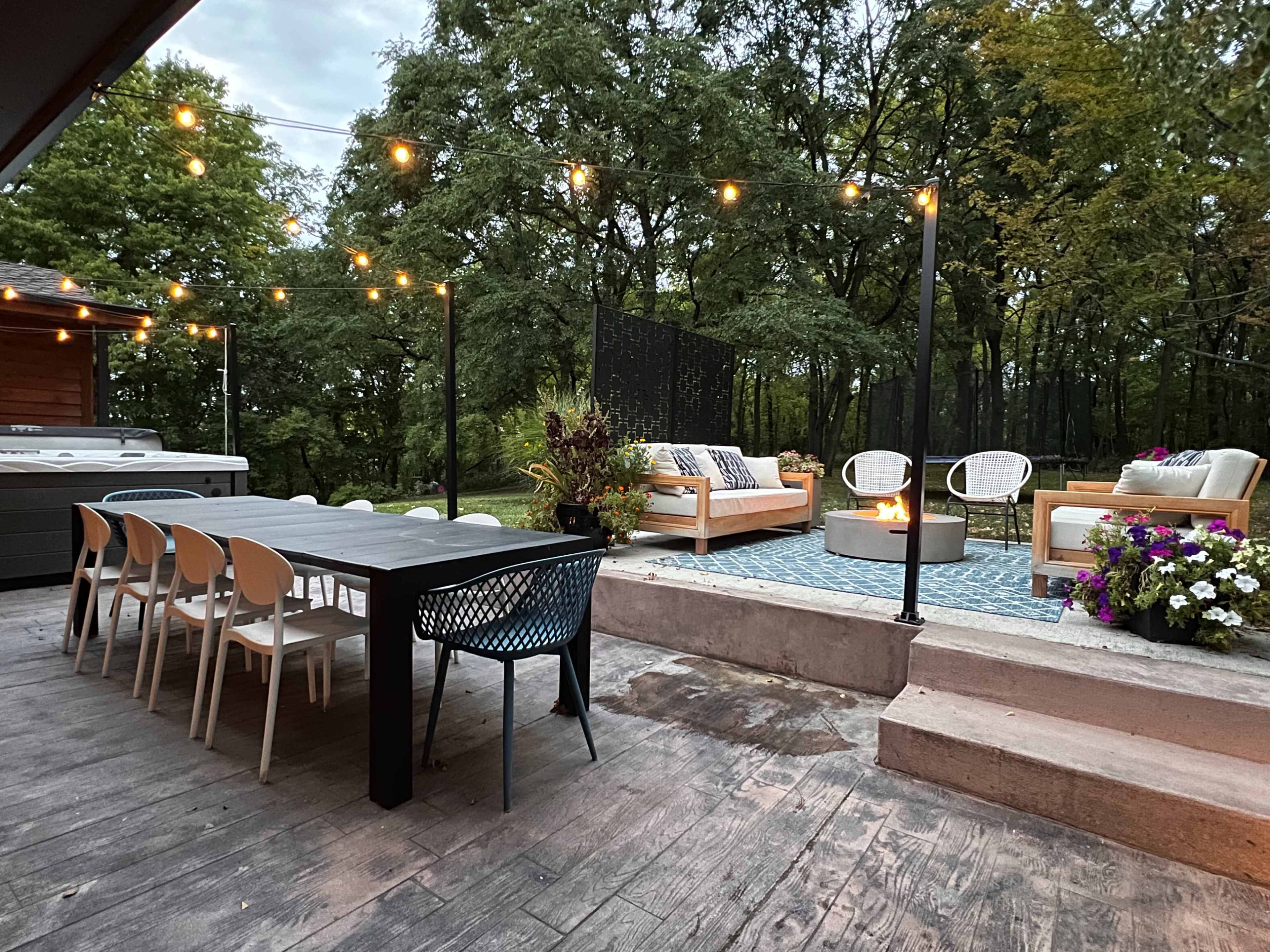 A backyard patio area with a long dining table, outdoor seating, a fire pit, and string lights overhead, surrounded by greenery.