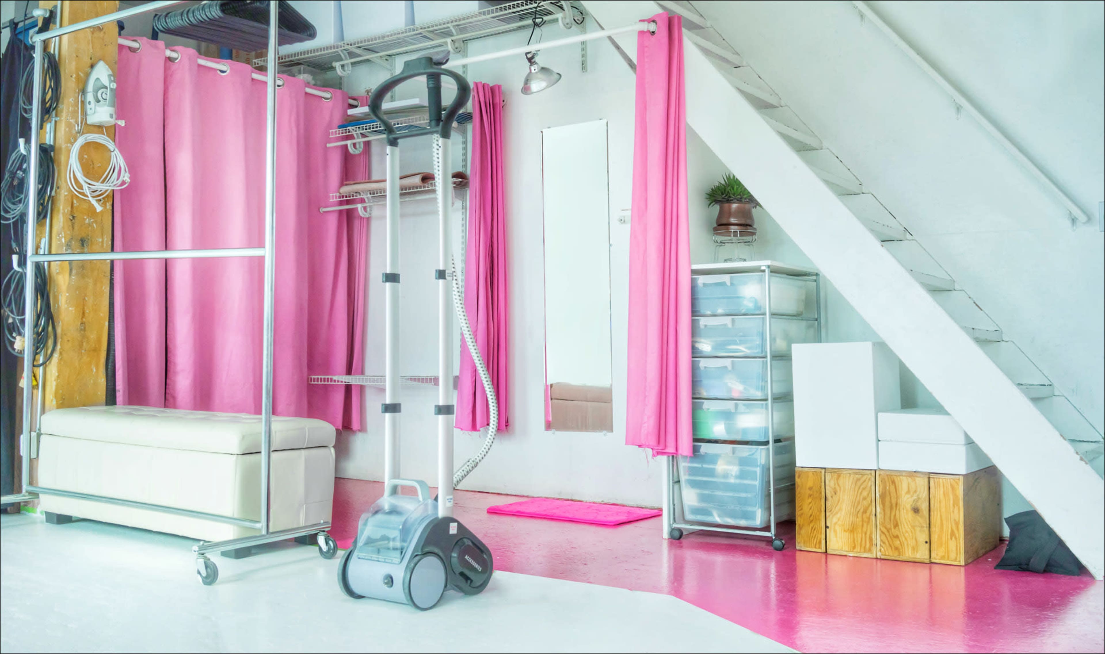 A bright, modern space featuring a white couch, pink curtains, a steamer, and storage units near a staircase.