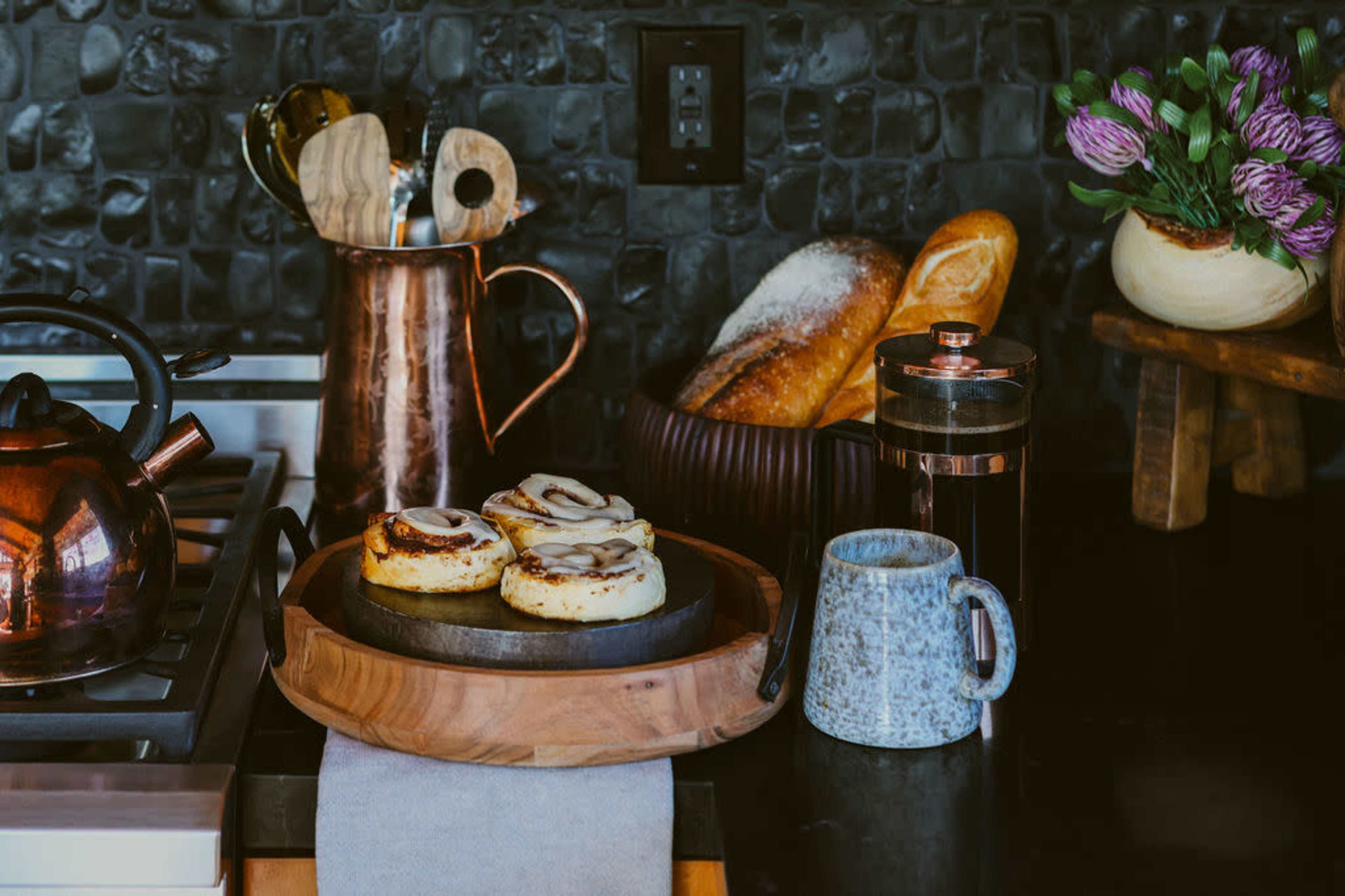 The image shows a kitchen countertop with a copper kettle, a wooden serving tray holding cinnamon rolls, a gray mug, and a French press, along with a basket of bread and a vase of purple flowers in the background.