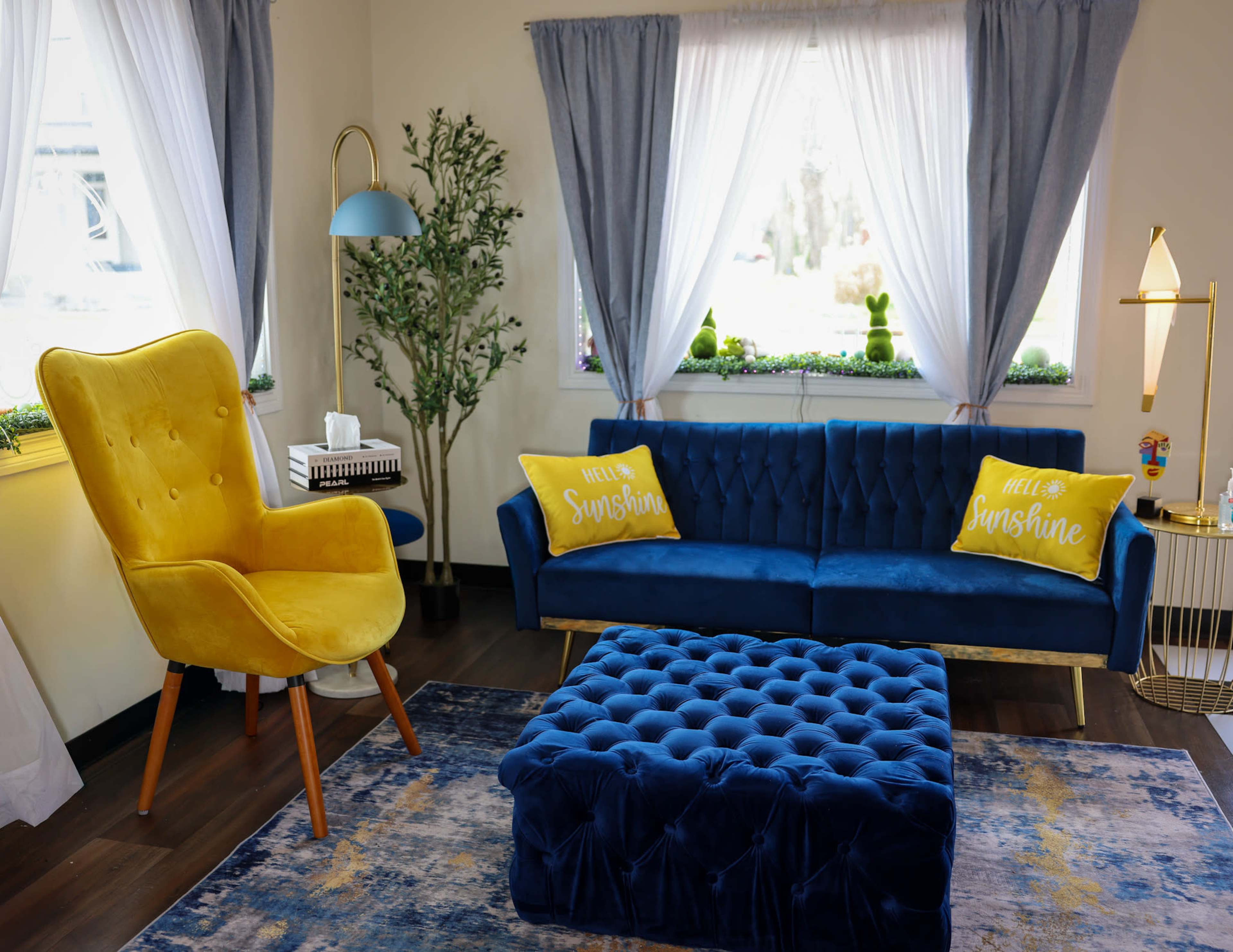 The image shows a brightly lit living room featuring a blue sofa with yellow "Hello Sunshine" pillows, a yellow accent chair, and a tufted blue ottoman on a patterned rug.