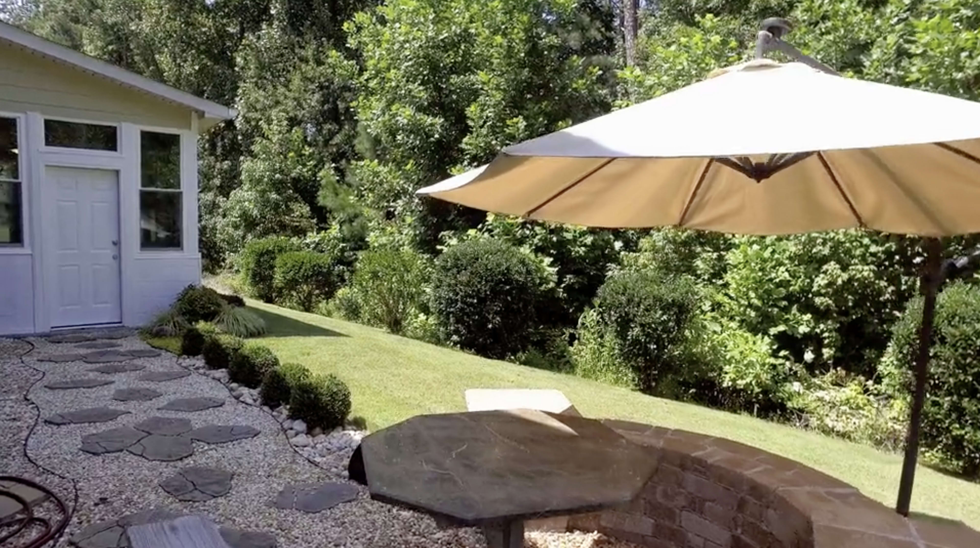A sunlit backyard with a stone pathway leading to a shaded seating area beneath a large umbrella and surrounded by lush greenery.