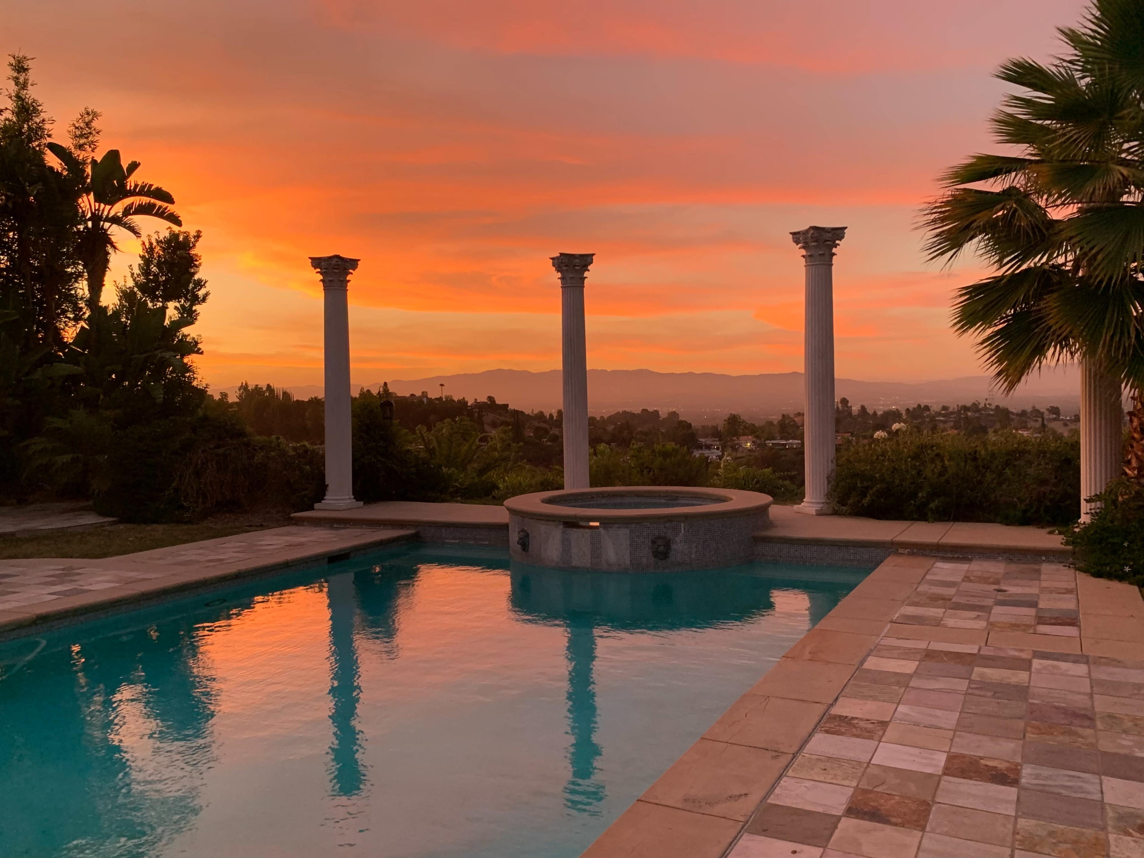 The scene shows a swimming pool with a stone patio, surrounded by tall columns and palm trees, against a sunset sky filled with pink and orange hues.