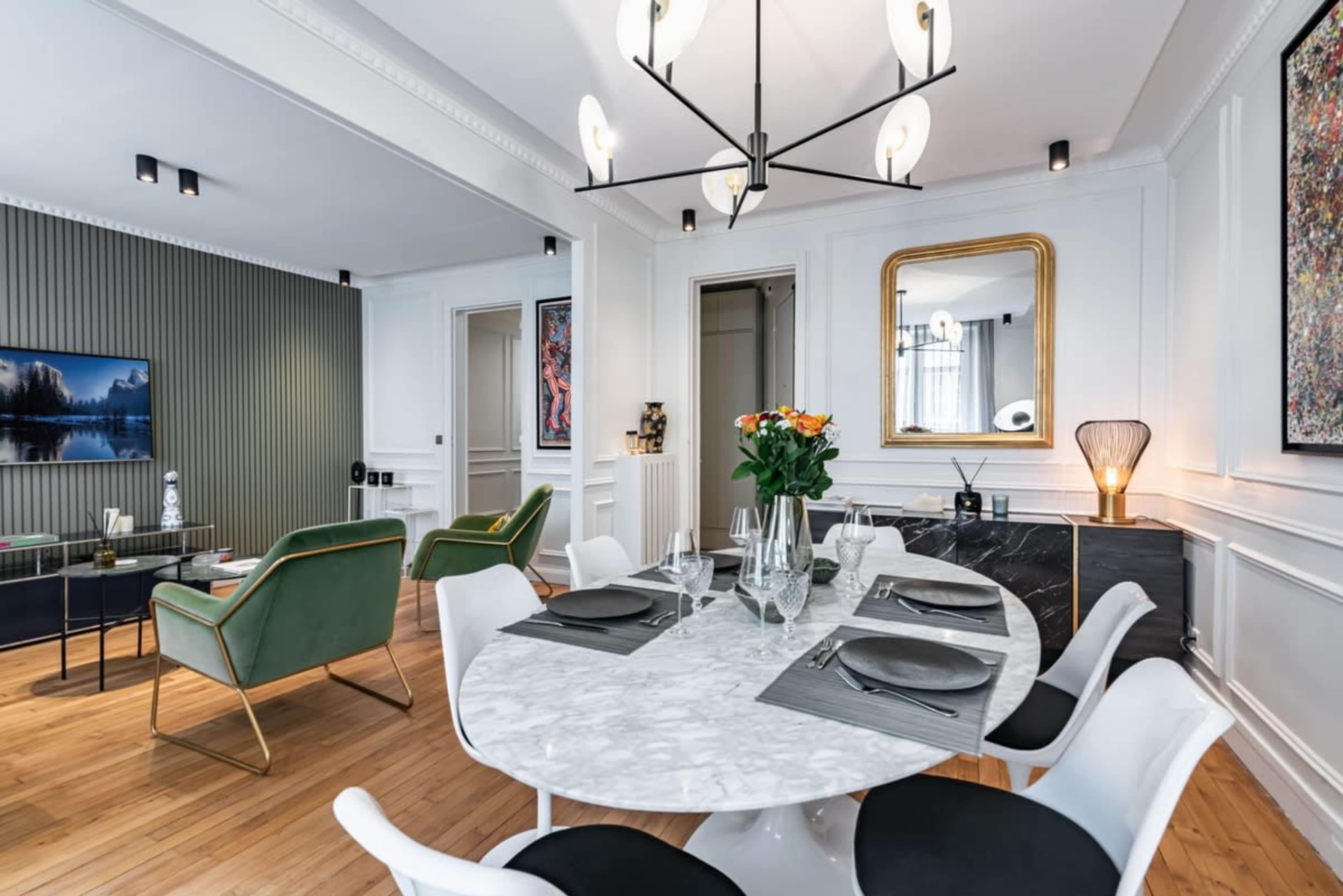 A circular marble dining table is set with dishware in a modern dining room featuring green chairs, a television, and decorative artwork on the walls.