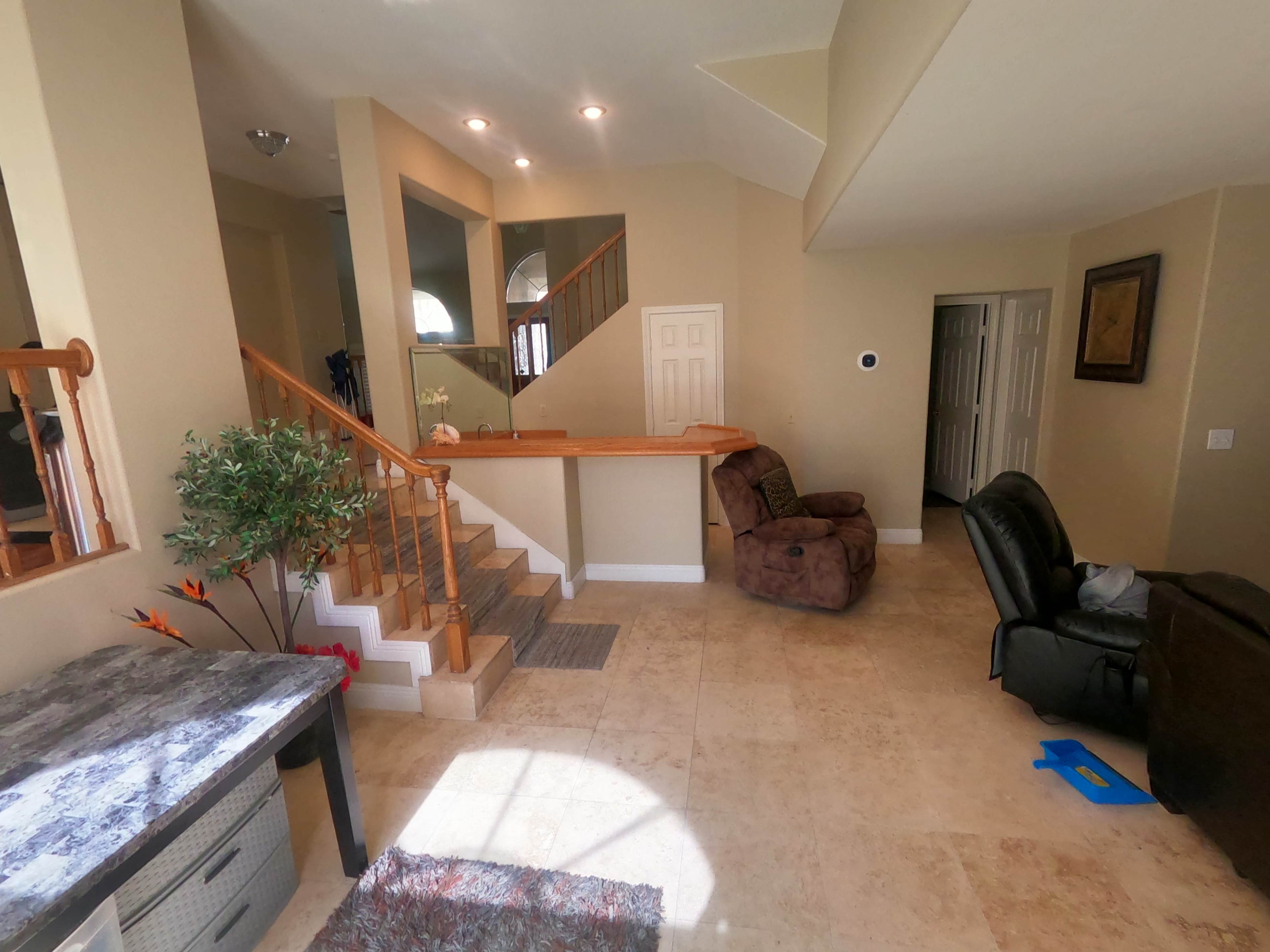 The image shows a spacious, open living area with natural stone flooring, a staircase, and a seating arrangement including a brown chair and a black recliner.