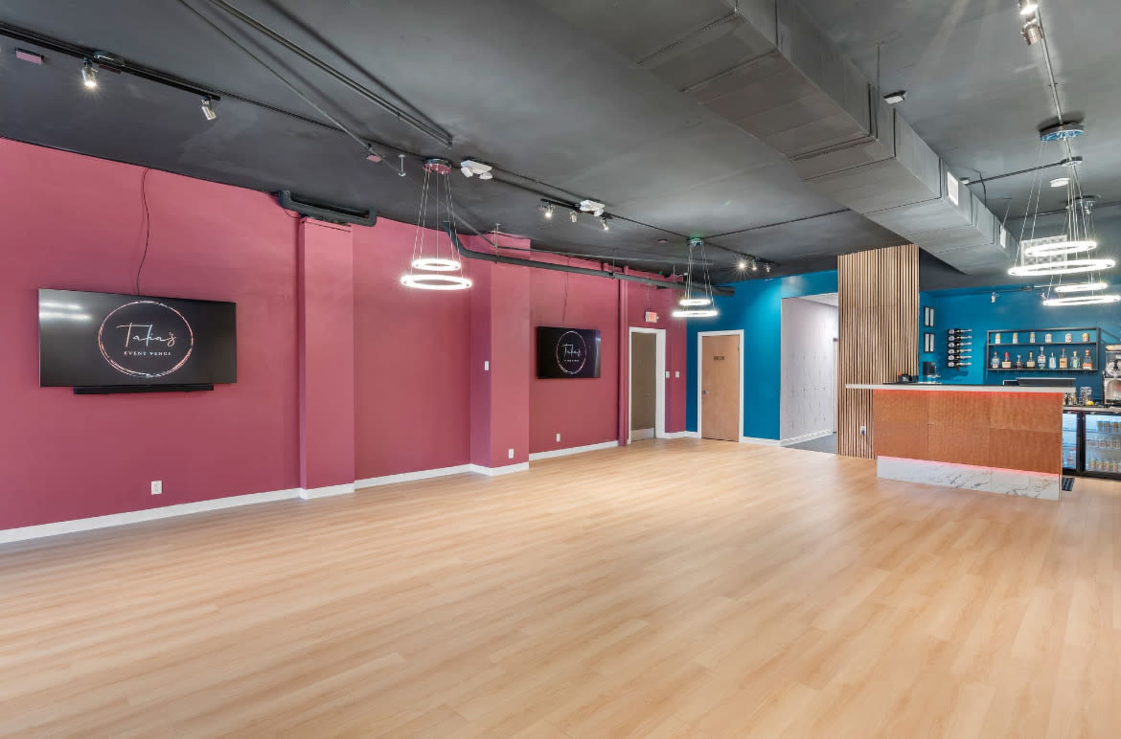 The image shows an empty interior space with pink and blue walls, wooden flooring, a bar area, and two wall-mounted screens.