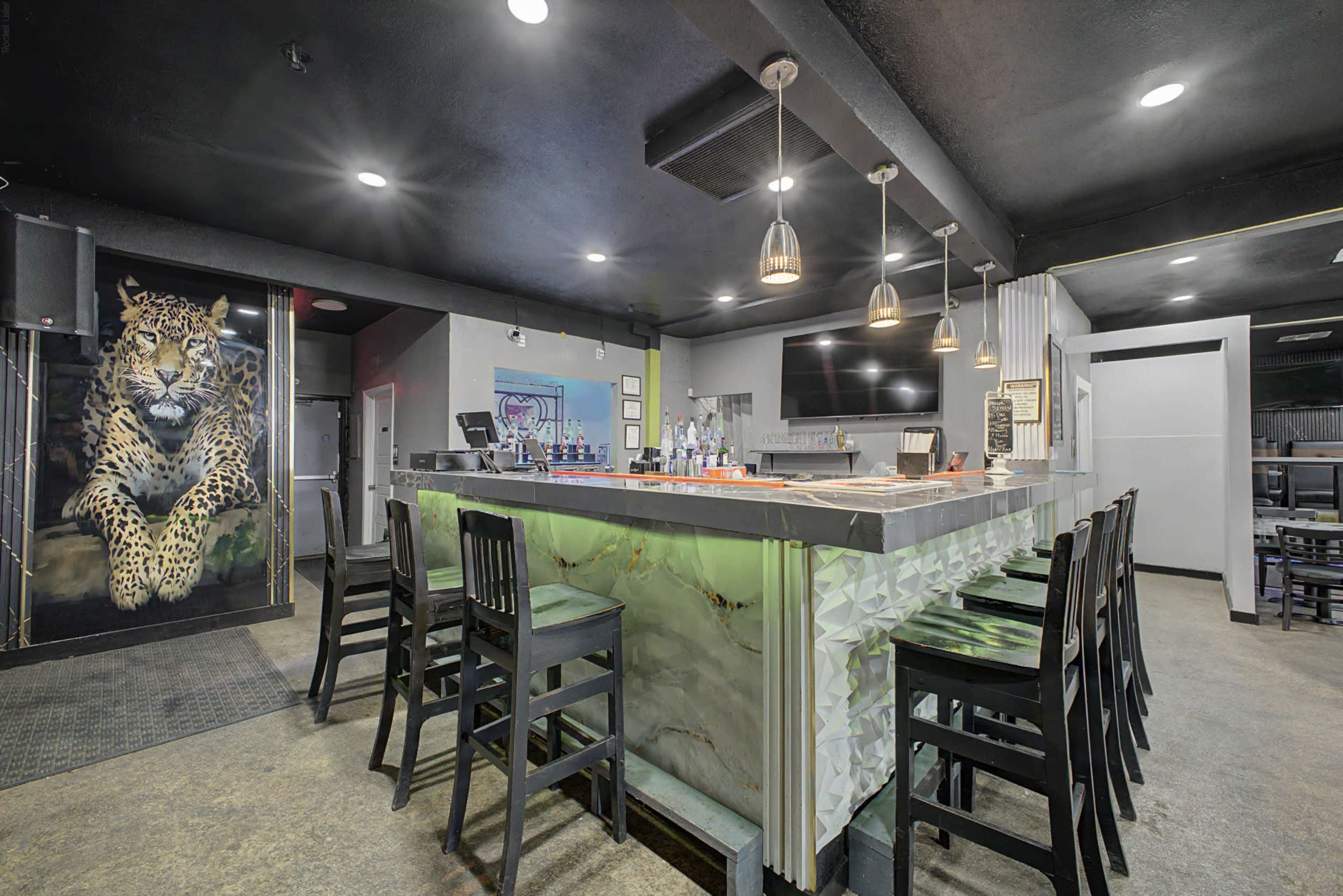 The image shows a modern bar area with a sleek countertop and high black stools, illuminated by pendant lights, featuring a large mural of a leopard on the wall.