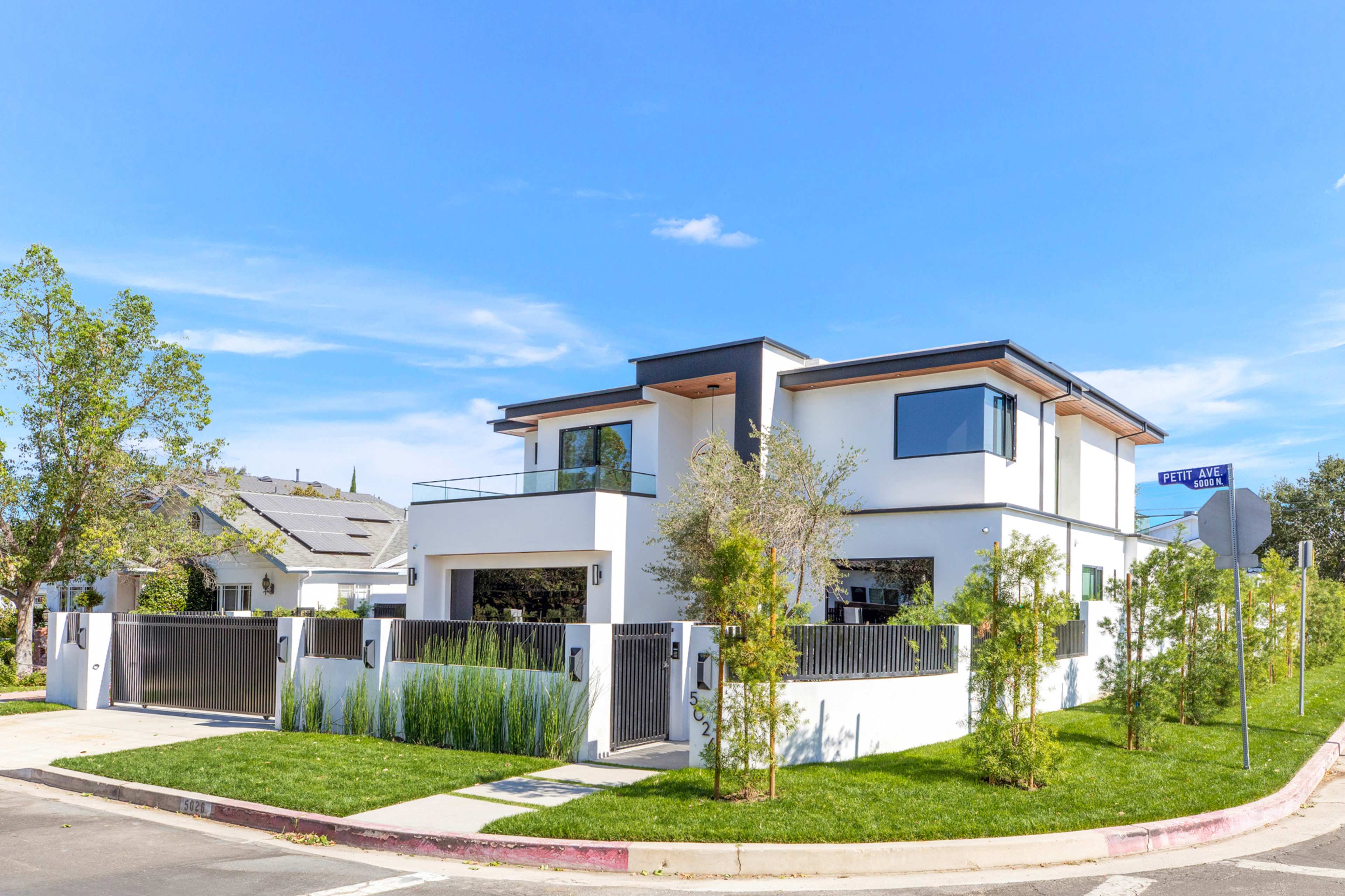 A modern two-story house with a flat roof and large windows is situated at the corner of a street, surrounded by greenery and a well-maintained lawn.
