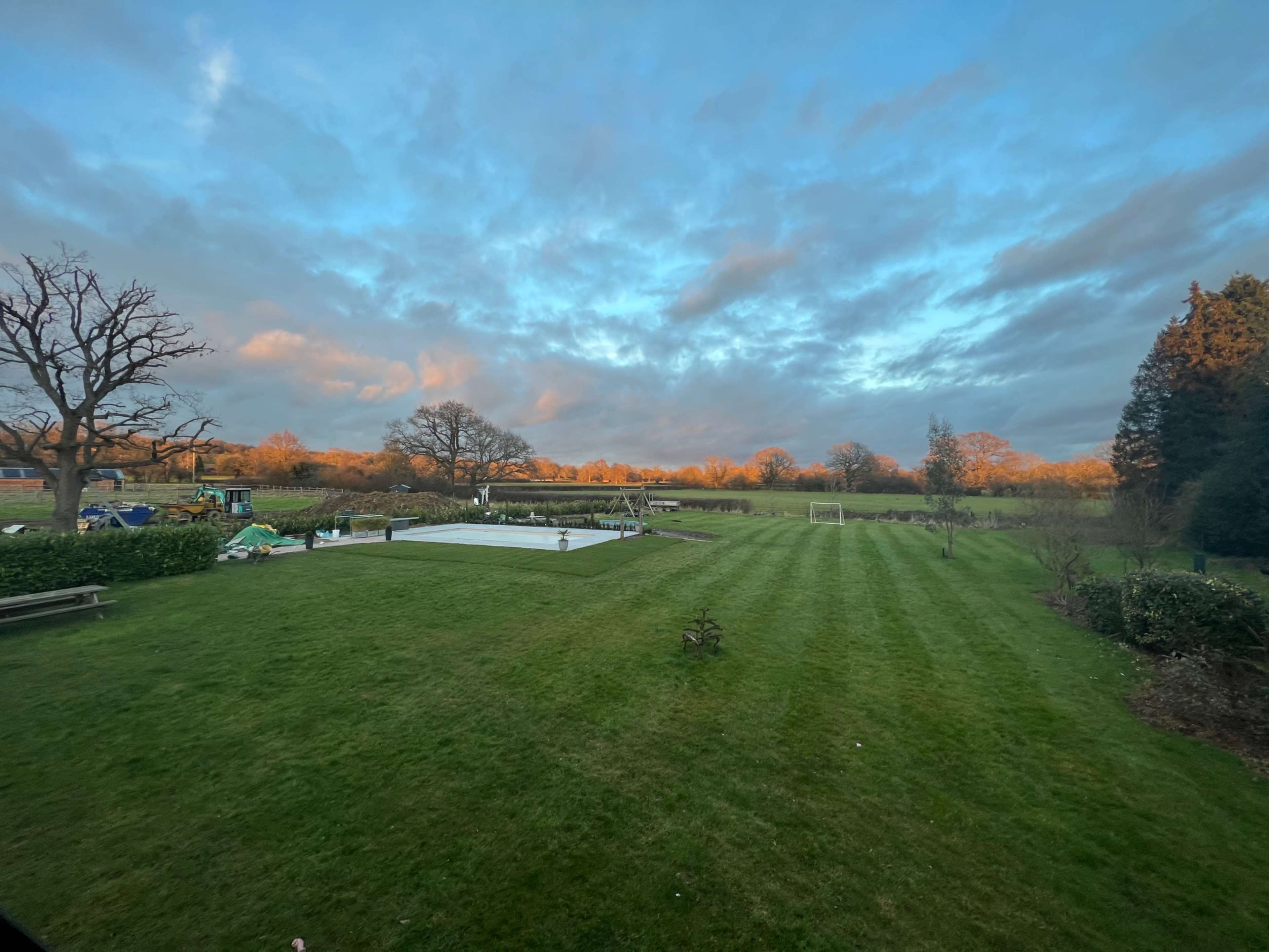 A large green lawn with a covered swimming pool and a soccer goal, set against a backdrop of trees and a cloudy sky.
