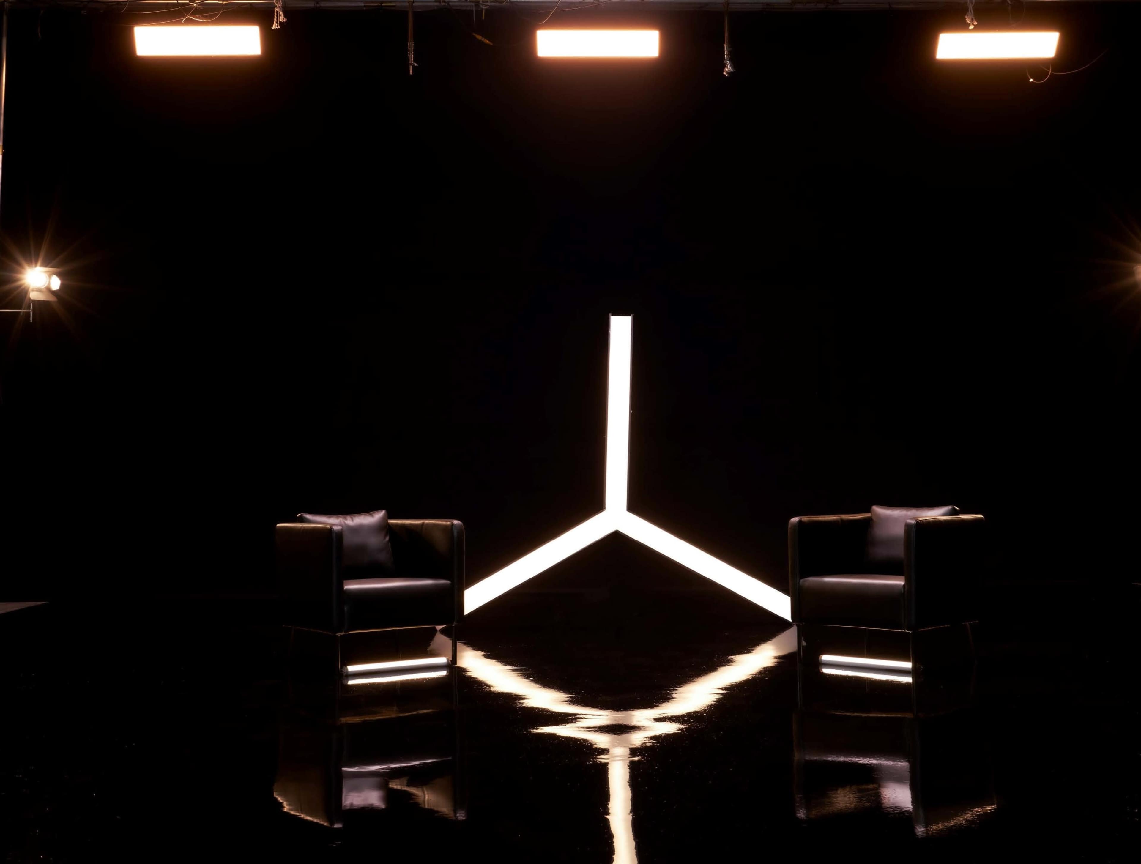 The image features a dark studio space with two black chairs facing a triangular light installation against a black backdrop.