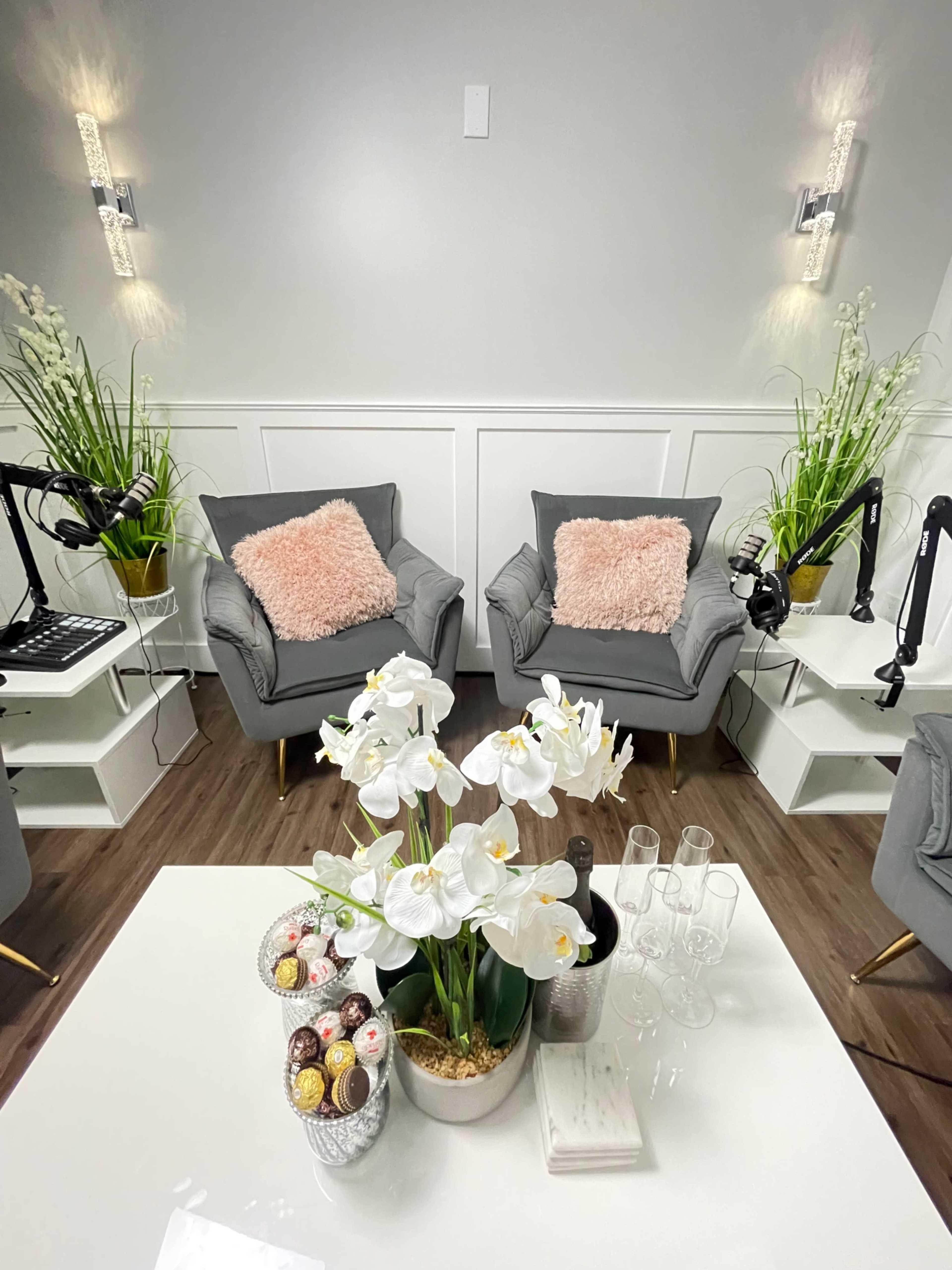 A modern lounge area features two gray chairs with pink cushions, a white coffee table adorned with flowers and decorative items, and green plants in the background.